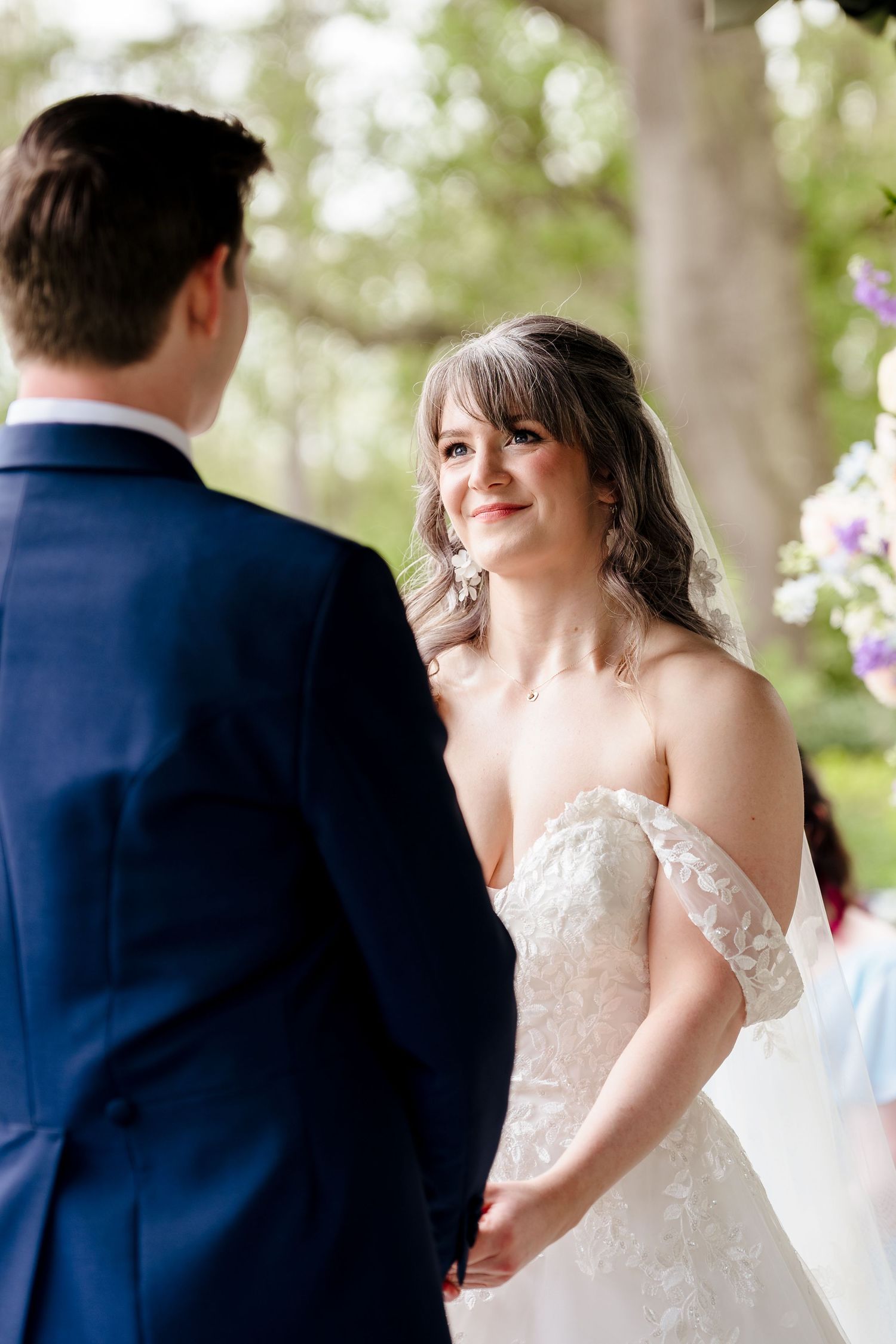 Smiling bride in off-shoulder lace gown holds grooms hands during outdoor wedding ceremony.