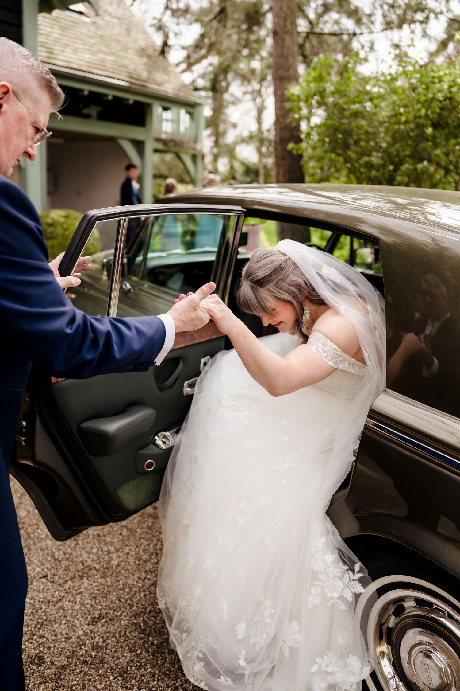 A bride in a lace gown being helped out of a vintage black car by a man in a navy suit.