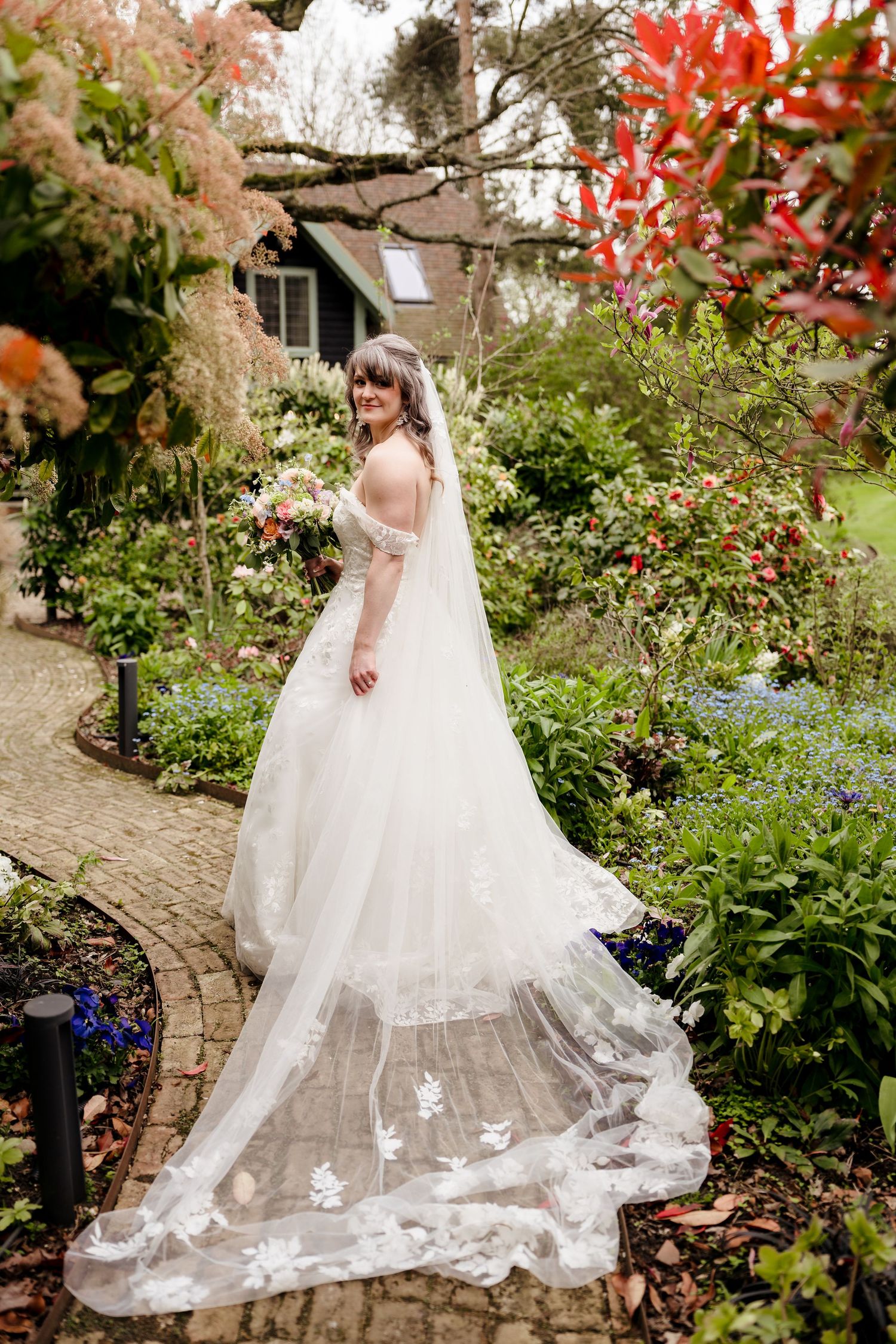 Bride in stunning lace gown with floral veil trails through lush garden pathway.