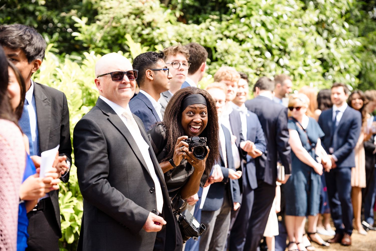 Diverse group of elegantly dressed guests at an outdoor event, with a smiling photographer capturing the moment.