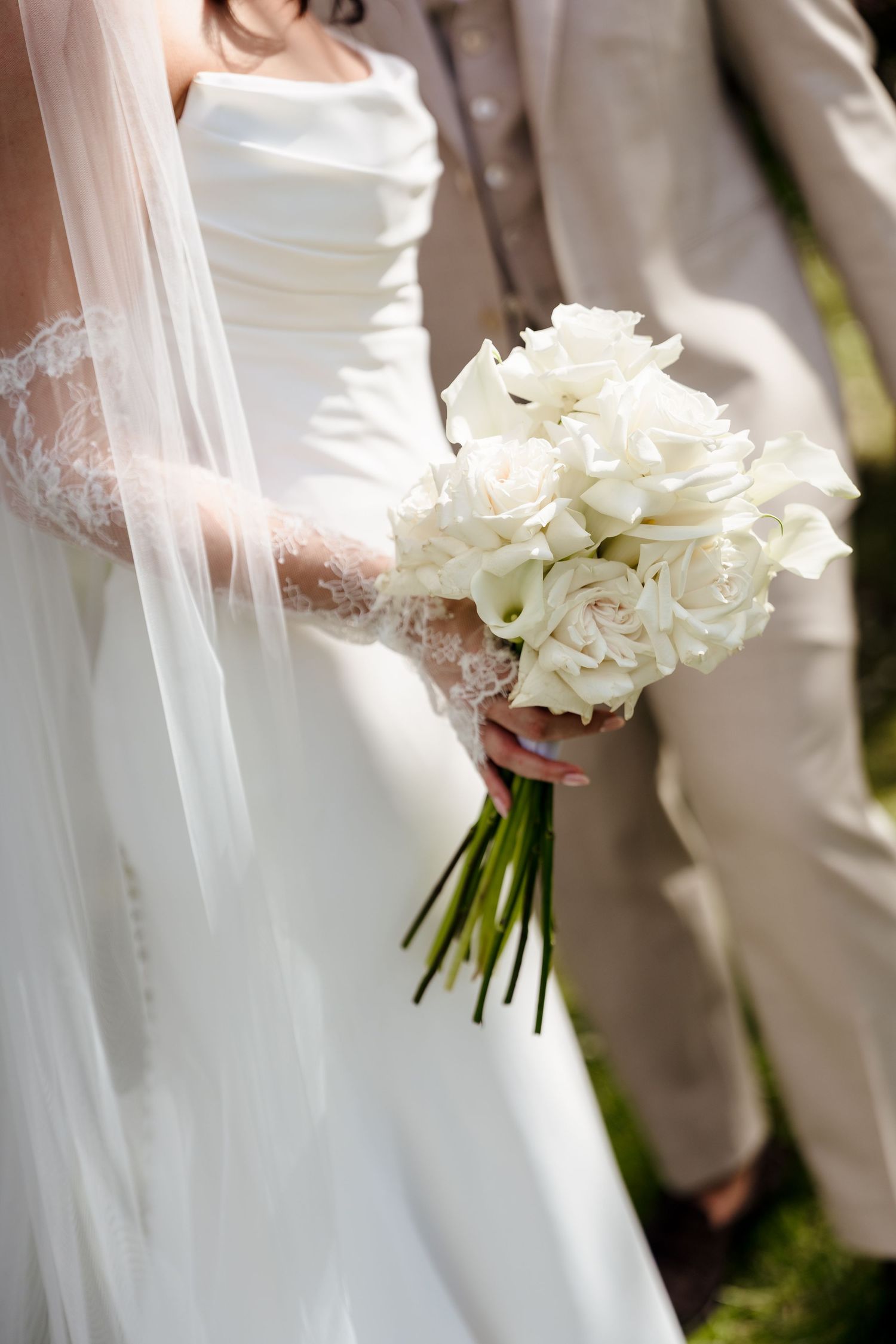 Bride in white gown and lace gloves holds classic bouquet of white roses with flowing veil.