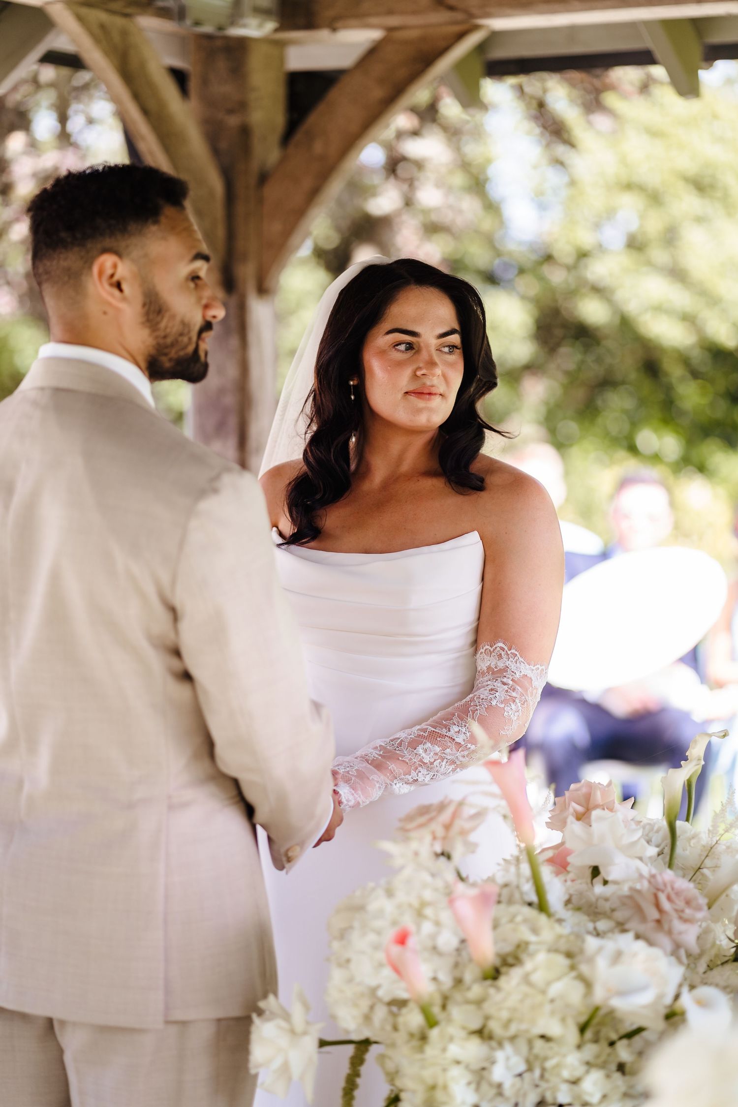 Bride in strapless white gown with lace gloves gazes at groom in beige suit during outdoor wedding ceremony.