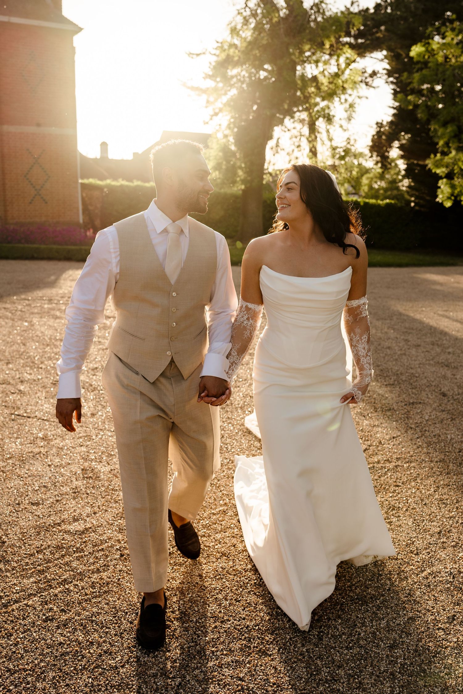 Bride and groom walk hand in hand at sunset, glowing backlight illuminating their elegant wedding attire.