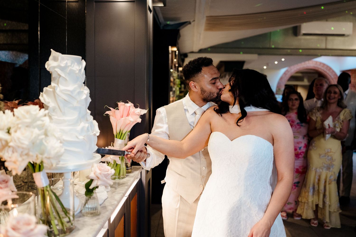 Bride and groom kissing while cutting white tiered wedding cake at reception.