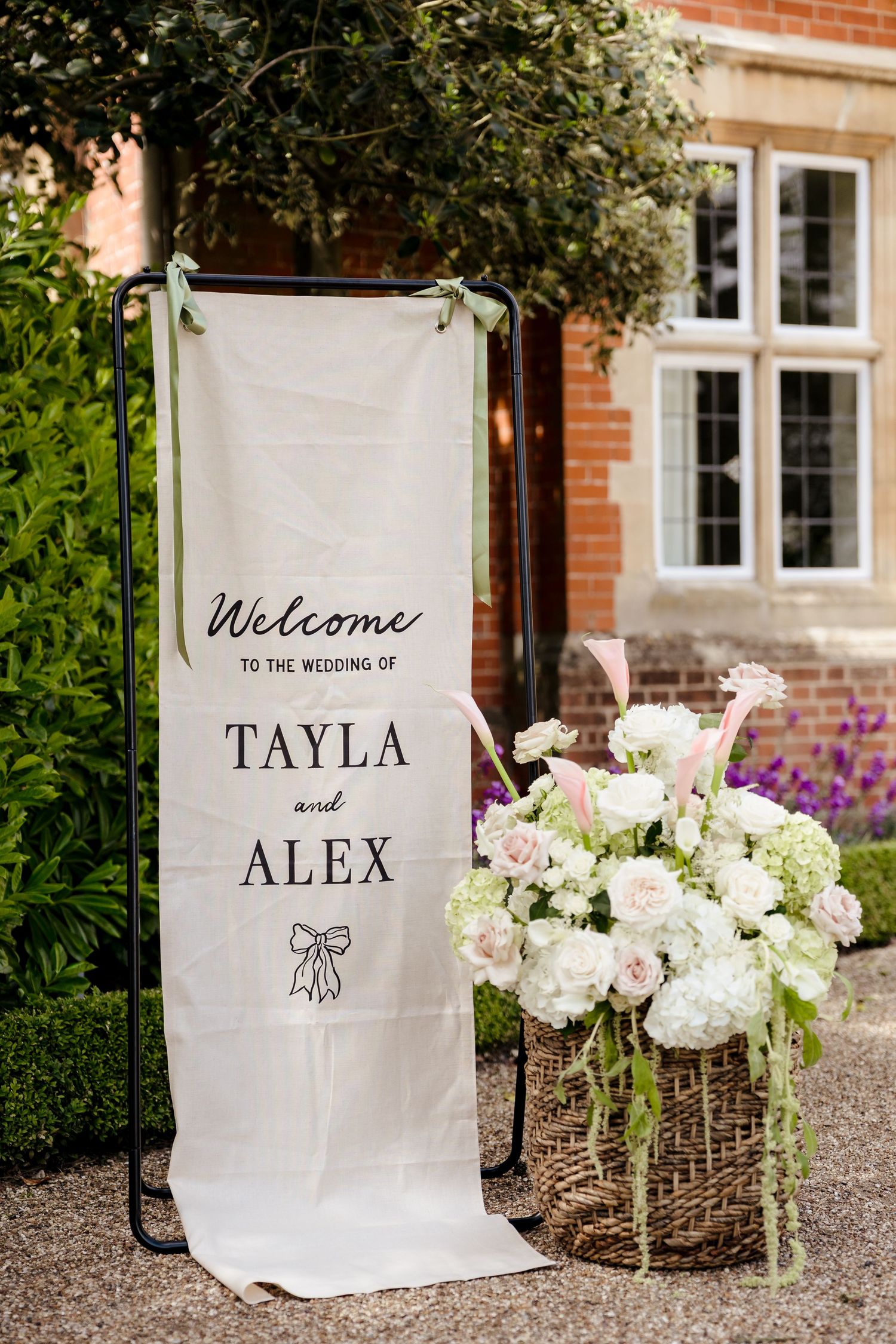 Elegant wedding welcome sign for Tayla and Alex with white floral arrangement in wicker basket outdoors.