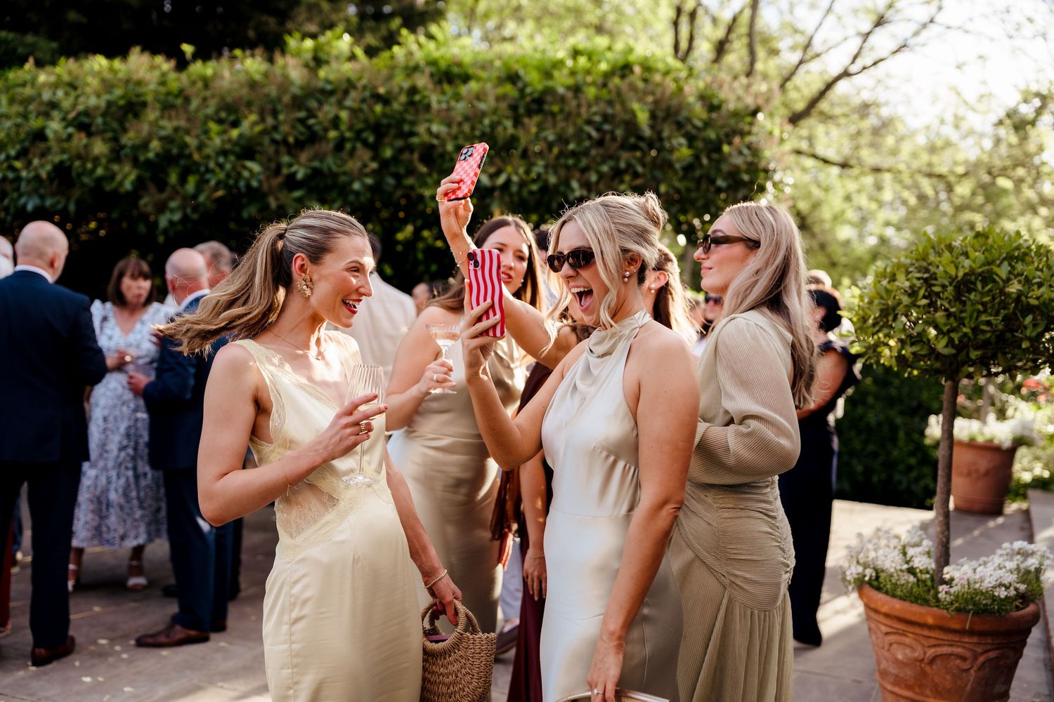 Bridesmaids in champagne satin dresses socializing outdoors at wedding reception on sunny day.