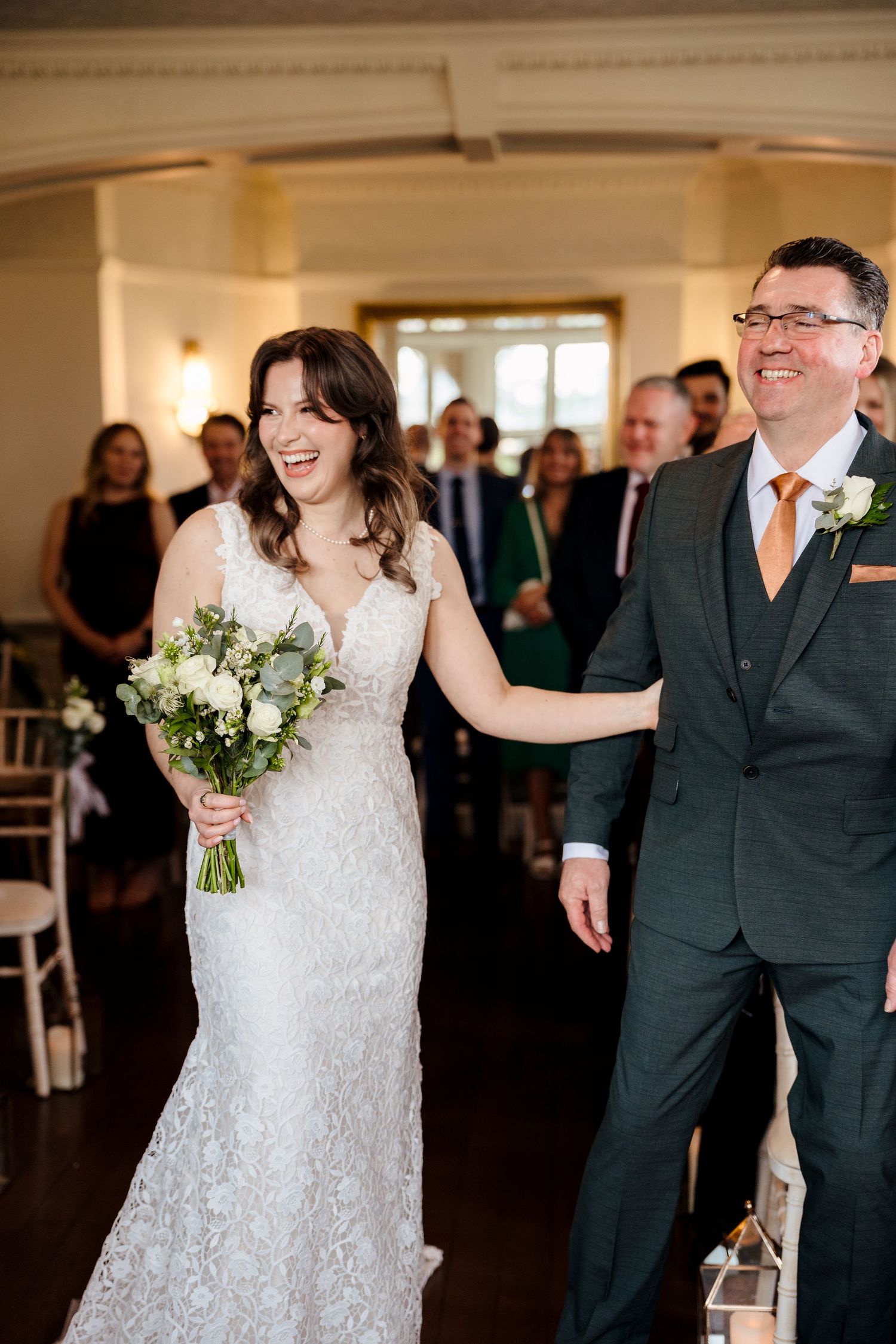 Joyful bride in lace gown holding bouquet walks down aisle with smiling groom in dark suit.