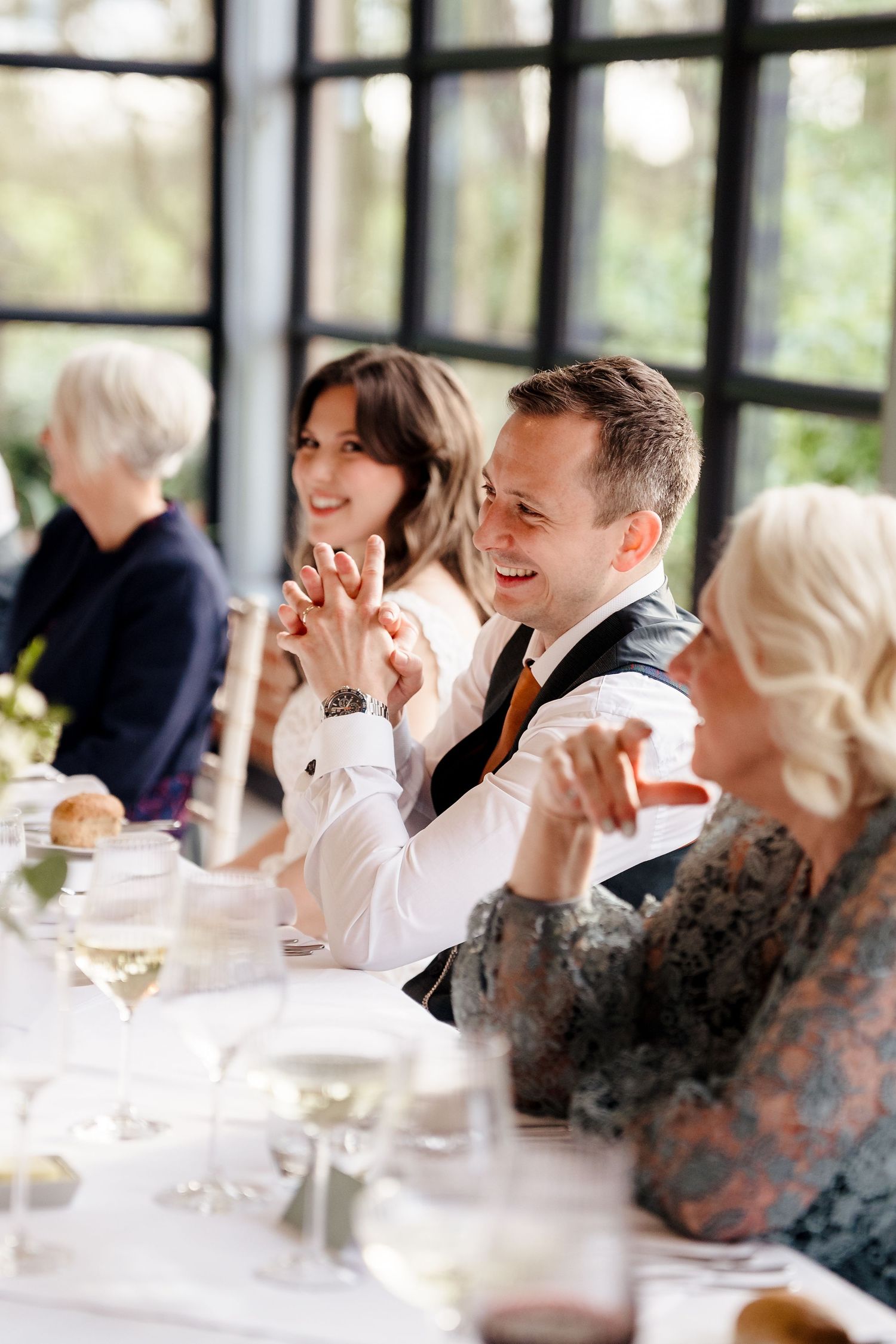 Wedding guests clap and smile at a reception dinner table with wine glasses and natural light background.