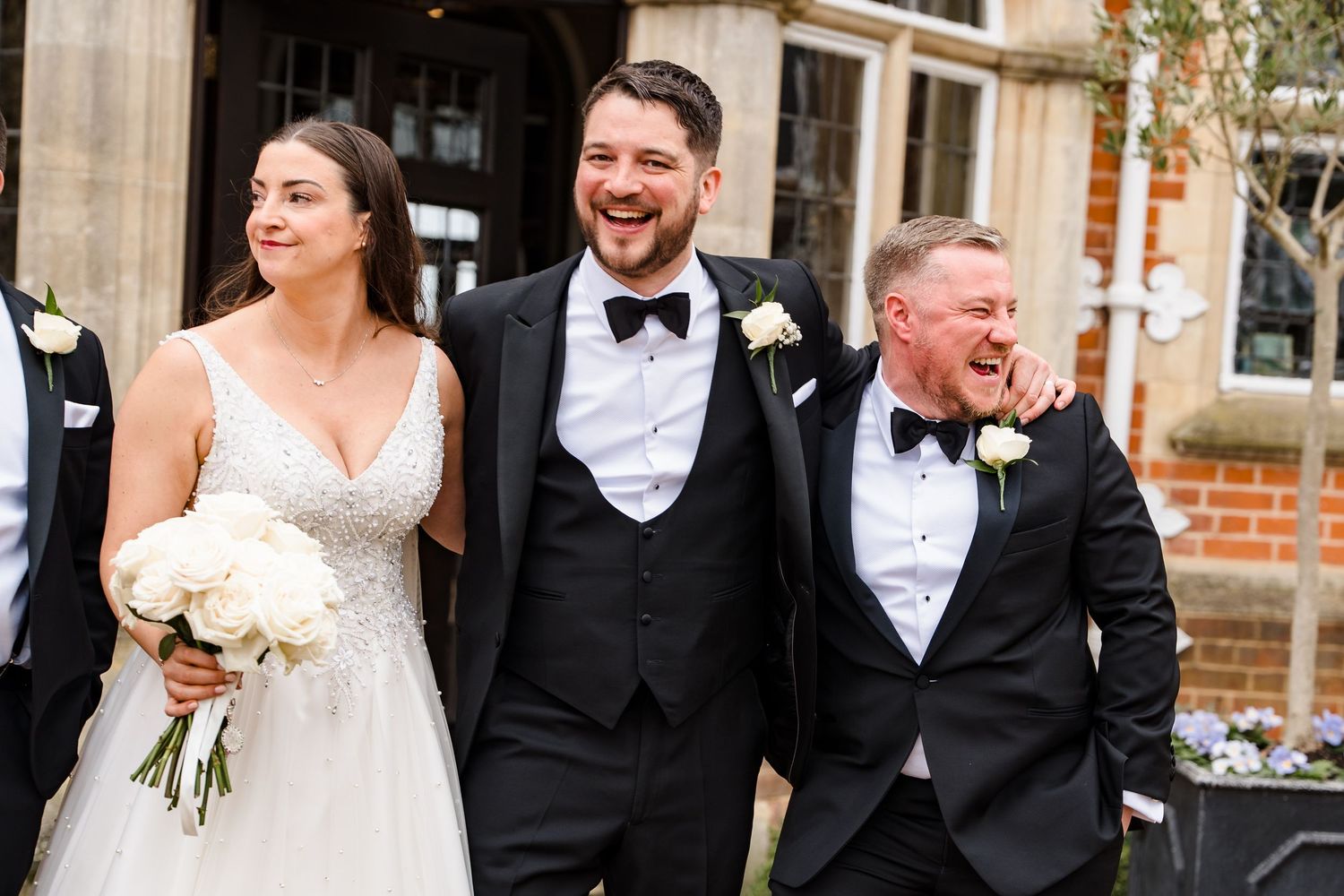 Bride in white gown and groom in tuxedo smiling with guest outside a brick building at a wedding.