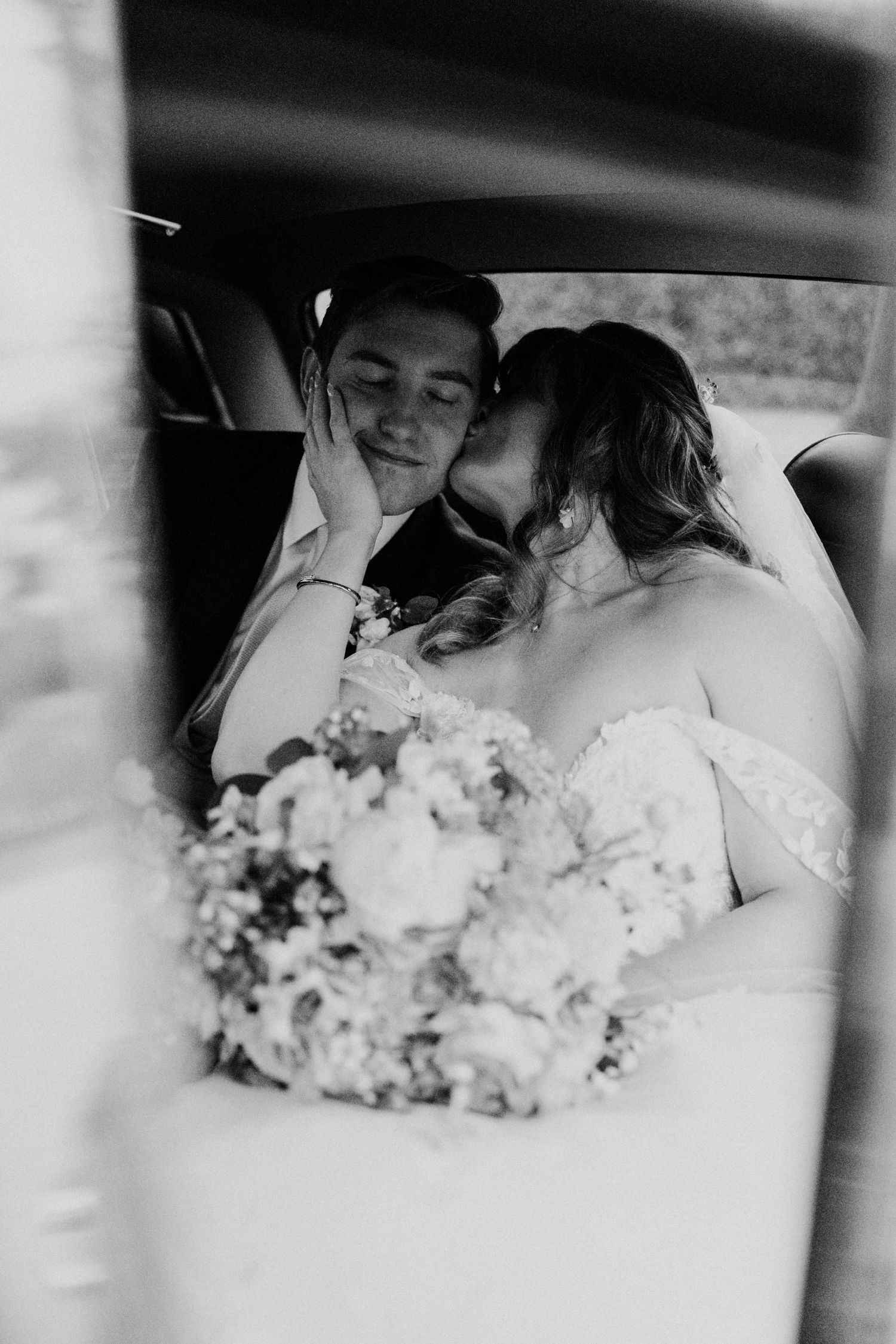 Black and white photo of bride kissing groom in car with wedding bouquet in foreground.
