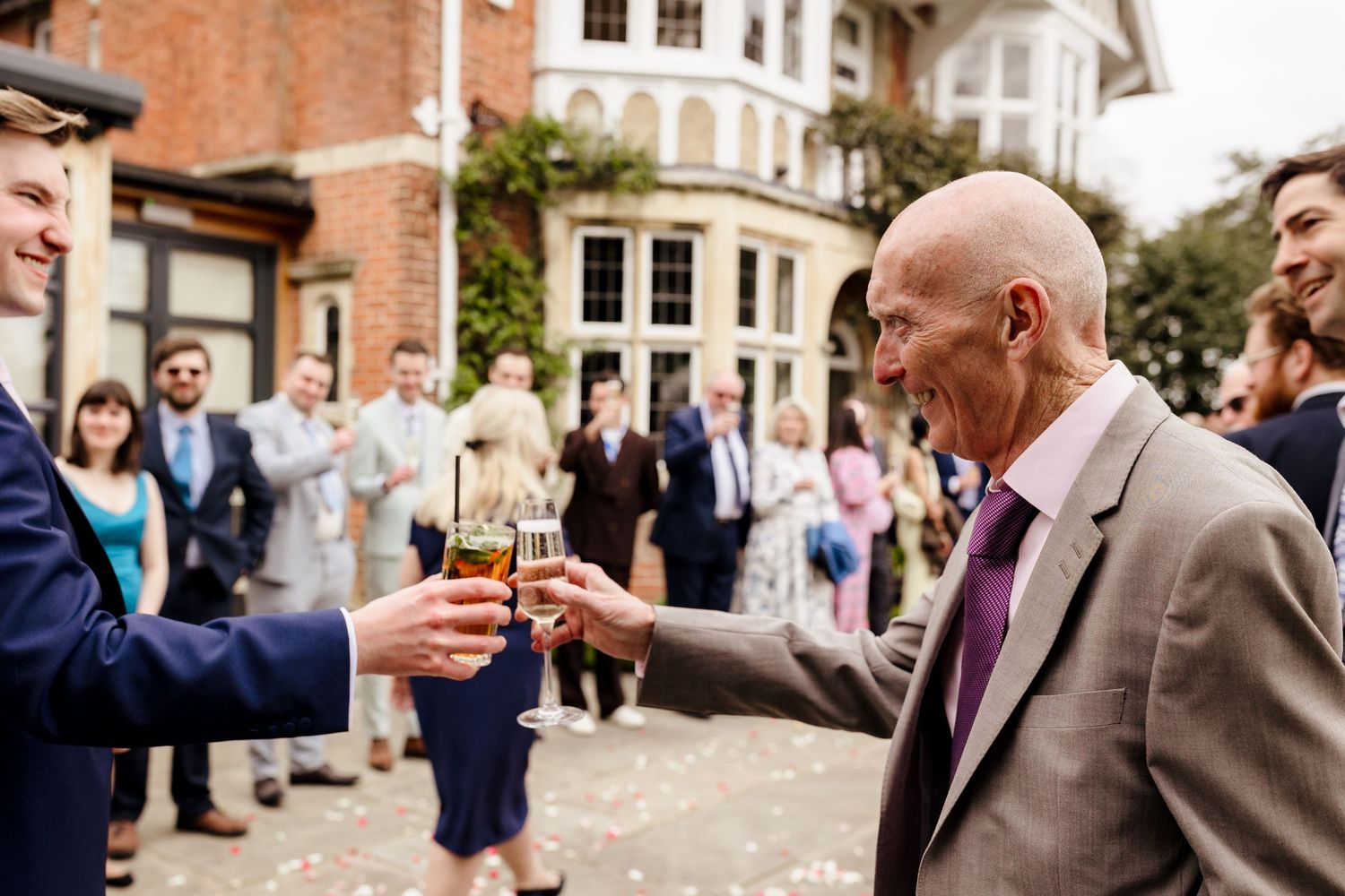 Wedding guests toasting with champagne glasses outside brick venue surrounded by confetti.