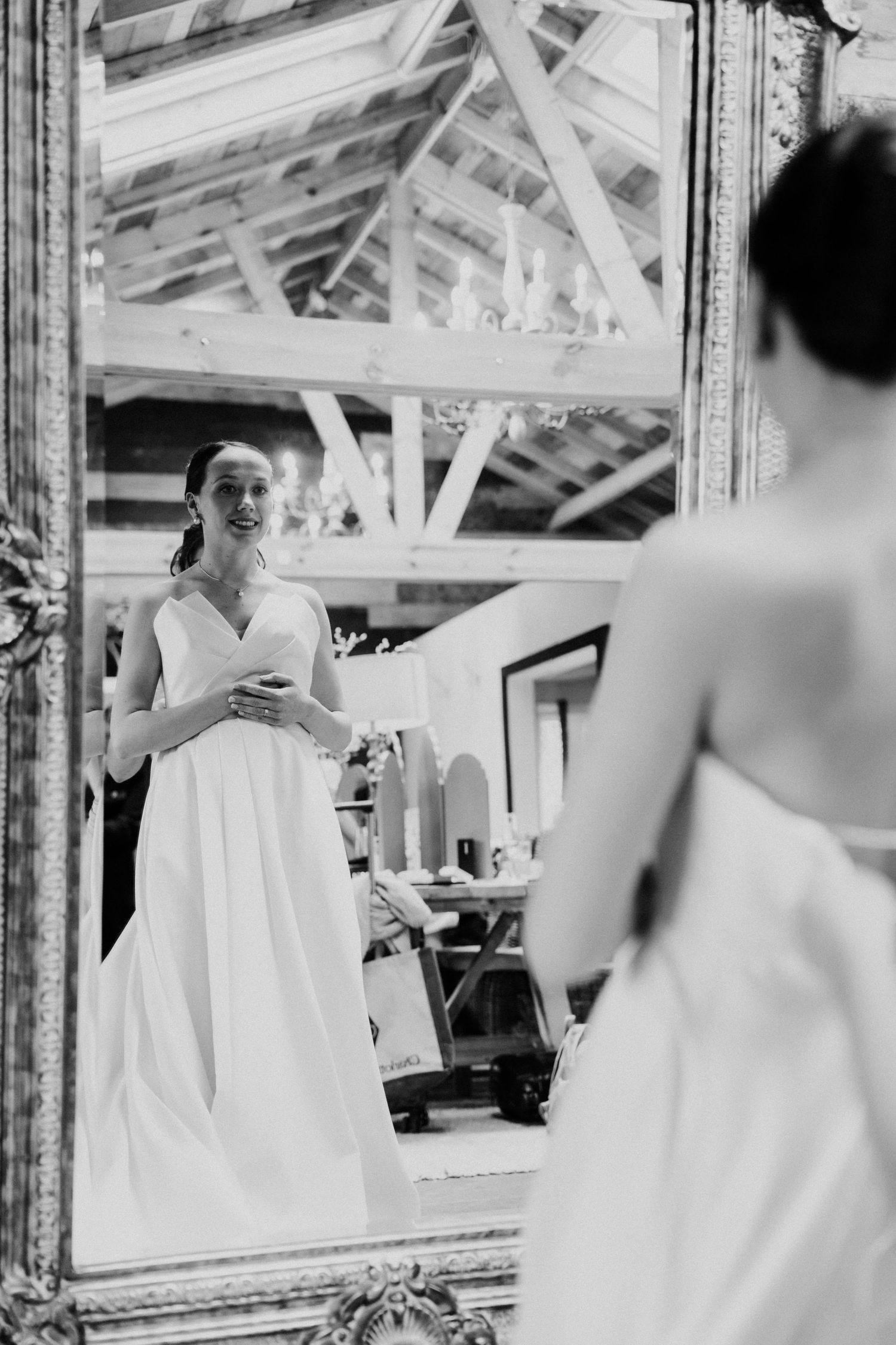 Black and white photo of a smiling bride in a white gown admiring herself in an ornate mirror inside a rustic venue.