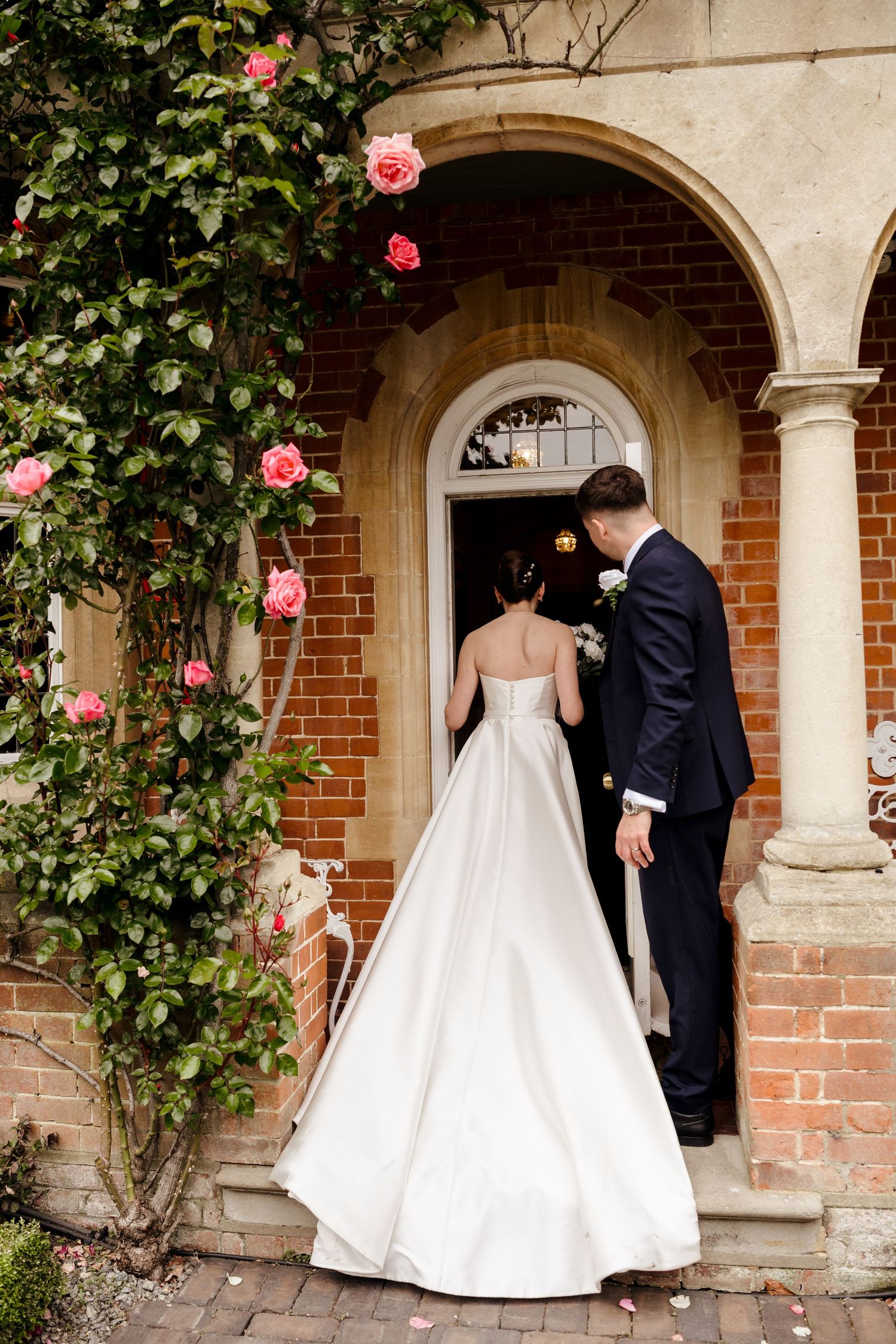 Bride and groom enter brick archway adorned with climbing pink roses at elegant English country house wedding venue.