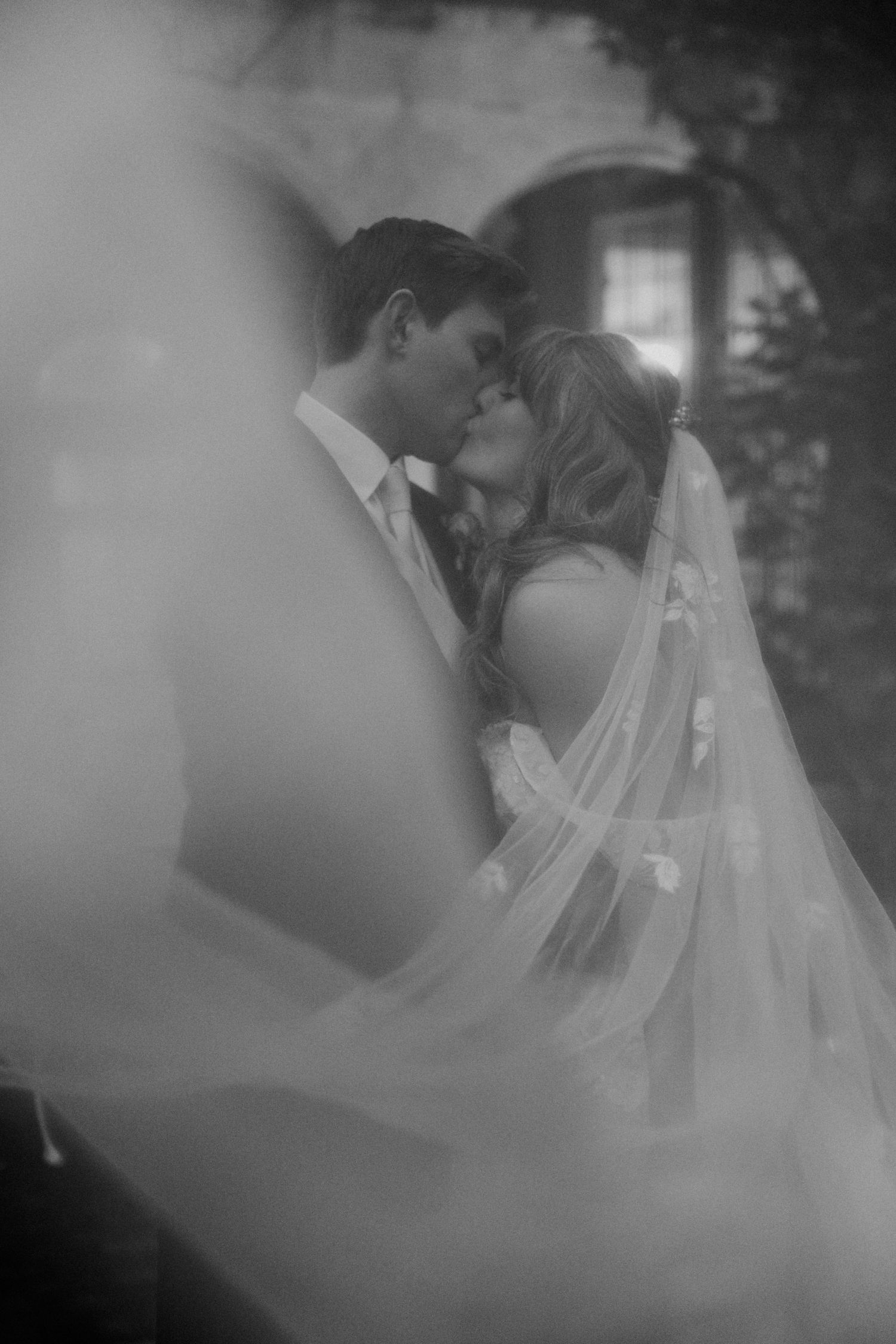 Romantic black and white photo of bride and groom kissing beneath a flowing veil at their wedding.