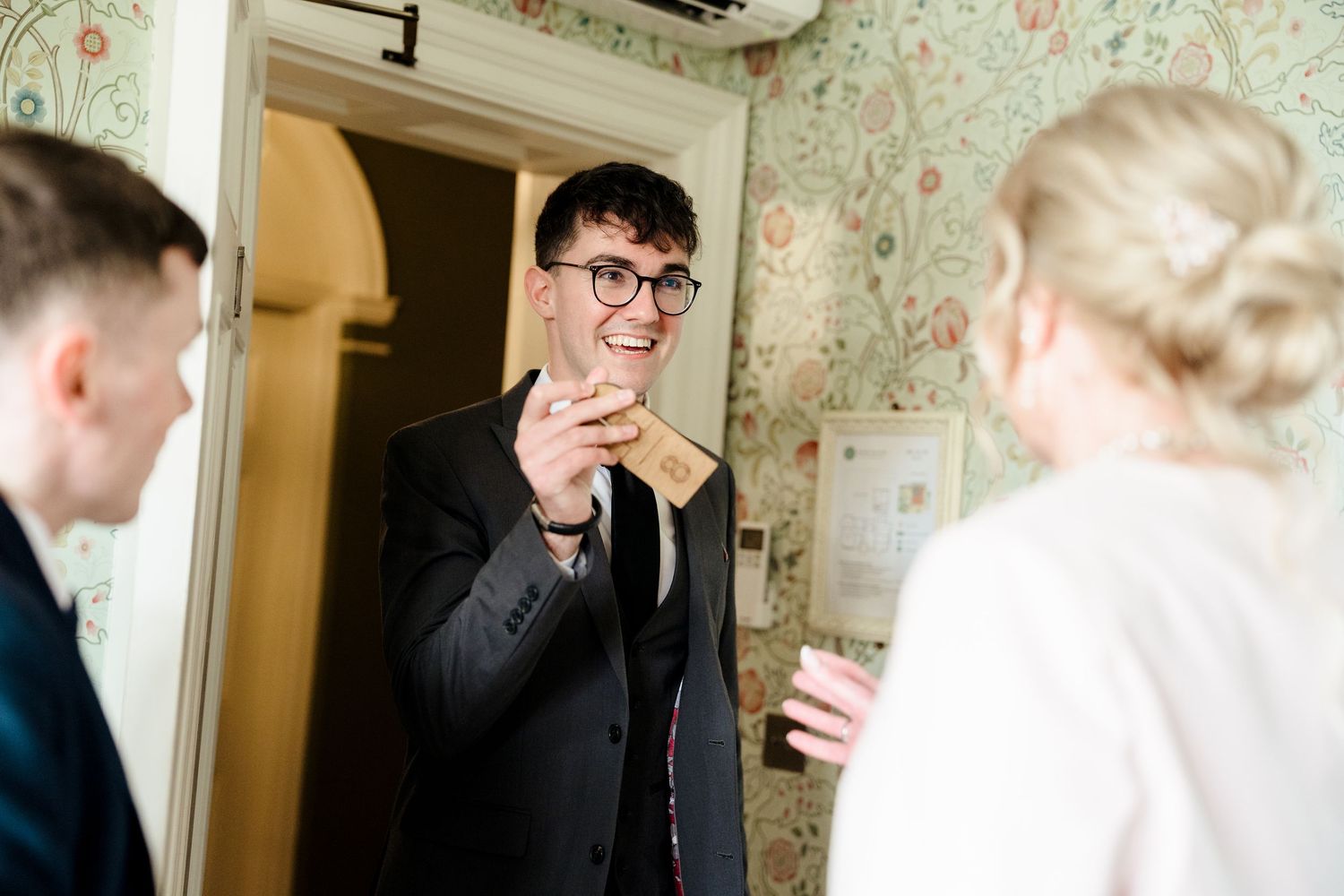 Smiling wedding guest in black suit adjusting tie and holding a gift inside a floral wallpapered room.