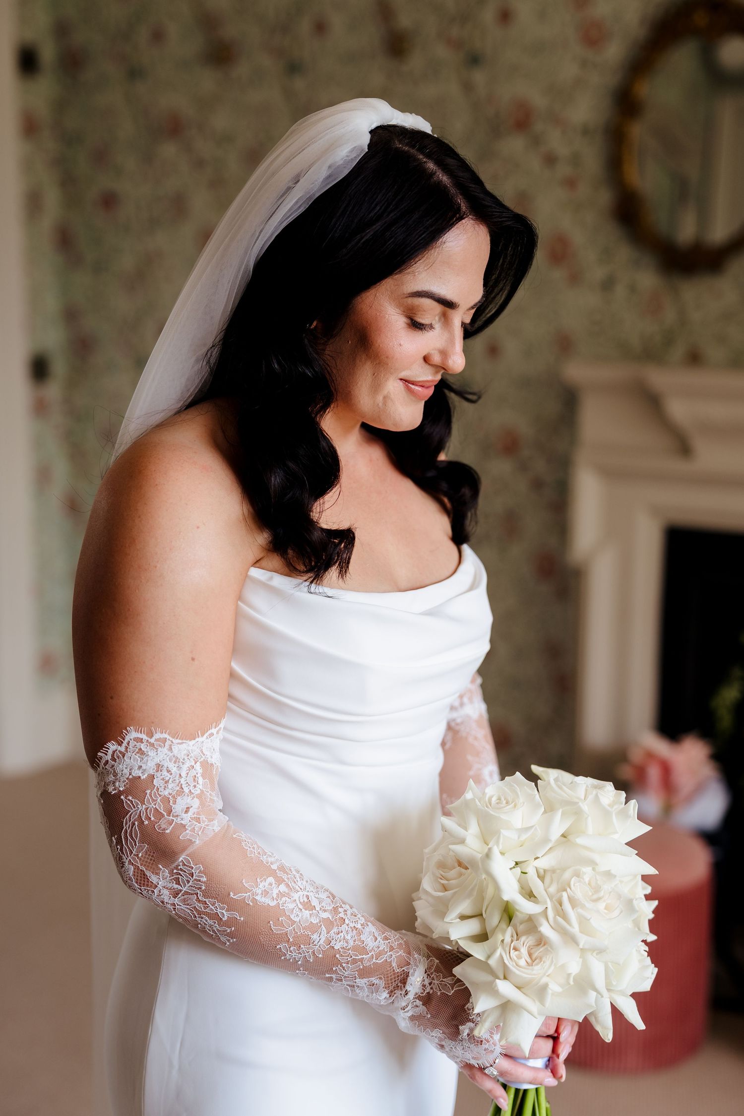 Bride in white gown with lace sleeves and veil holds white rose bouquet, smiling softly in elegant floral room.