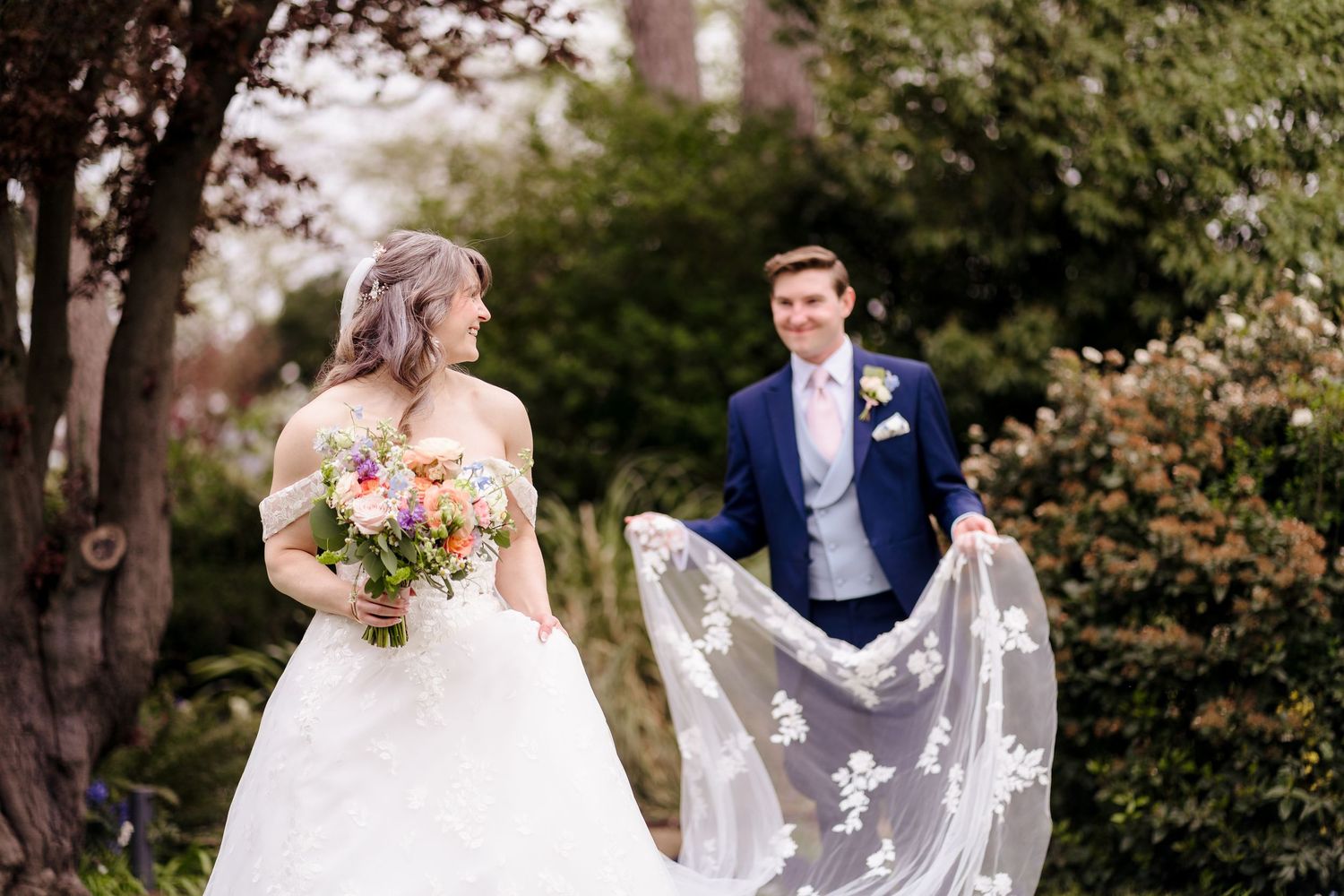Bride twirling lace veil while groom smiles in lush garden wedding portrait.