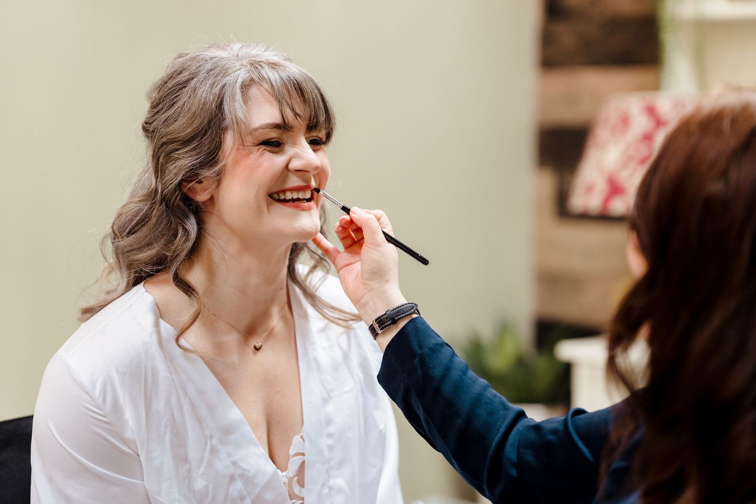 Makeup artist applying blush to a smiling woman with gray hair in a white blouse at a beauty salon.
