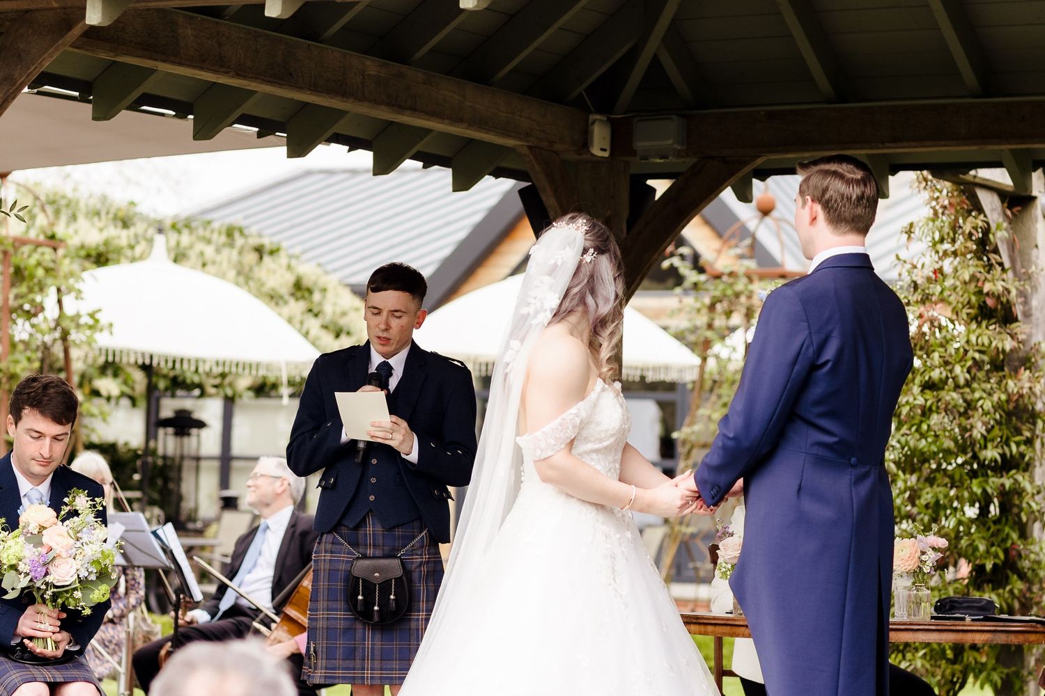 Officiant in tartan kilt reads vows as bride in white gown and groom in navy suit hold hands at outdoor wedding ceremony.