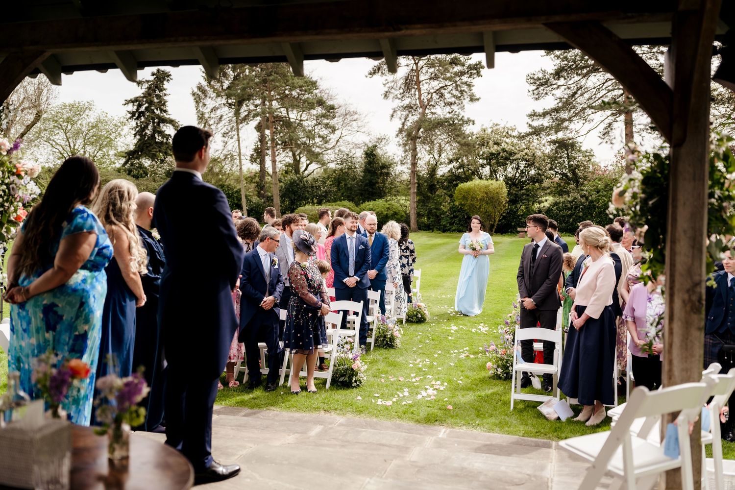 Outdoor wedding ceremony under a wooden pavilion with guests seated on white chairs in a lush garden.
