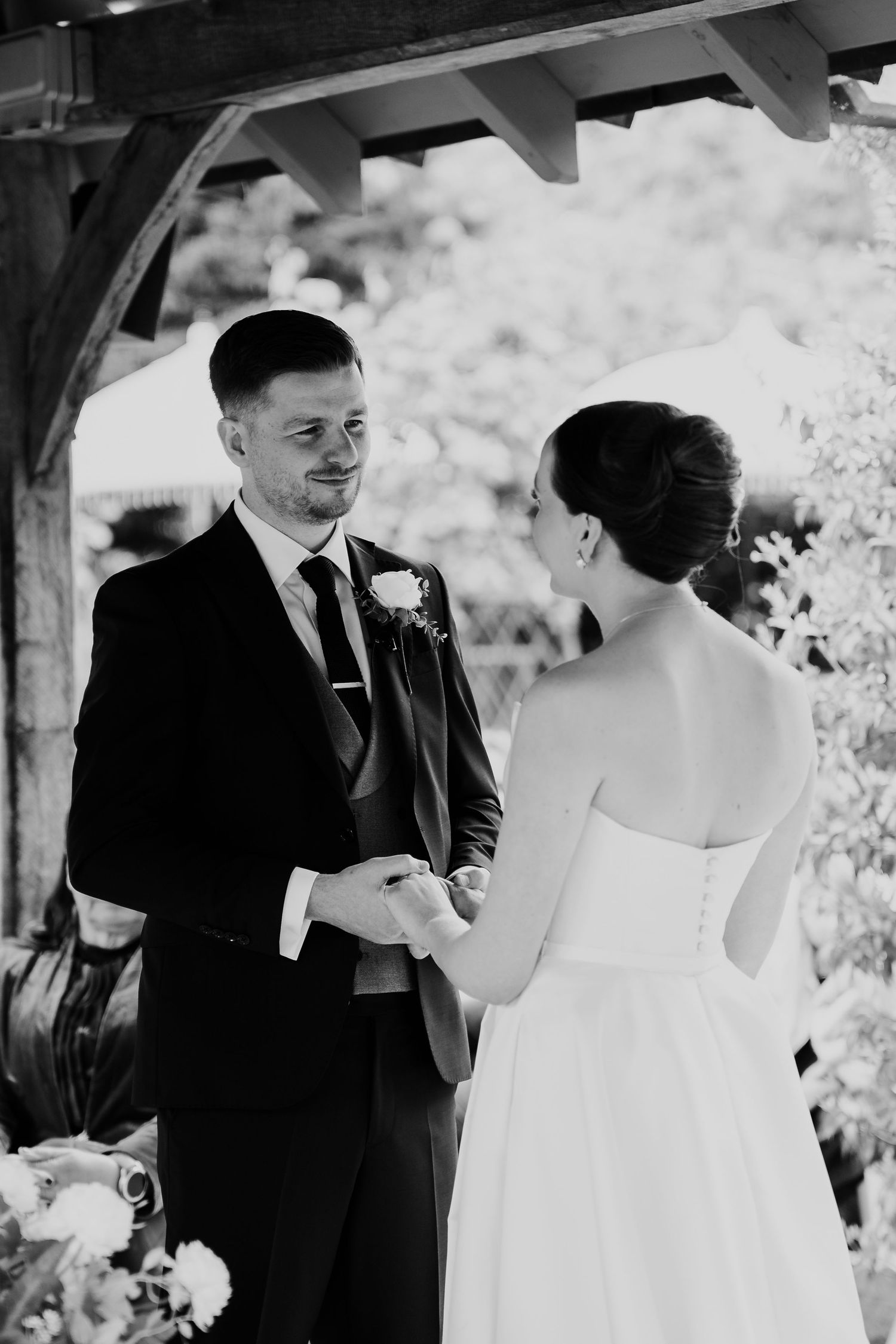 Black and white photo of groom smiling at bride while holding hands during outdoor wedding ceremony.