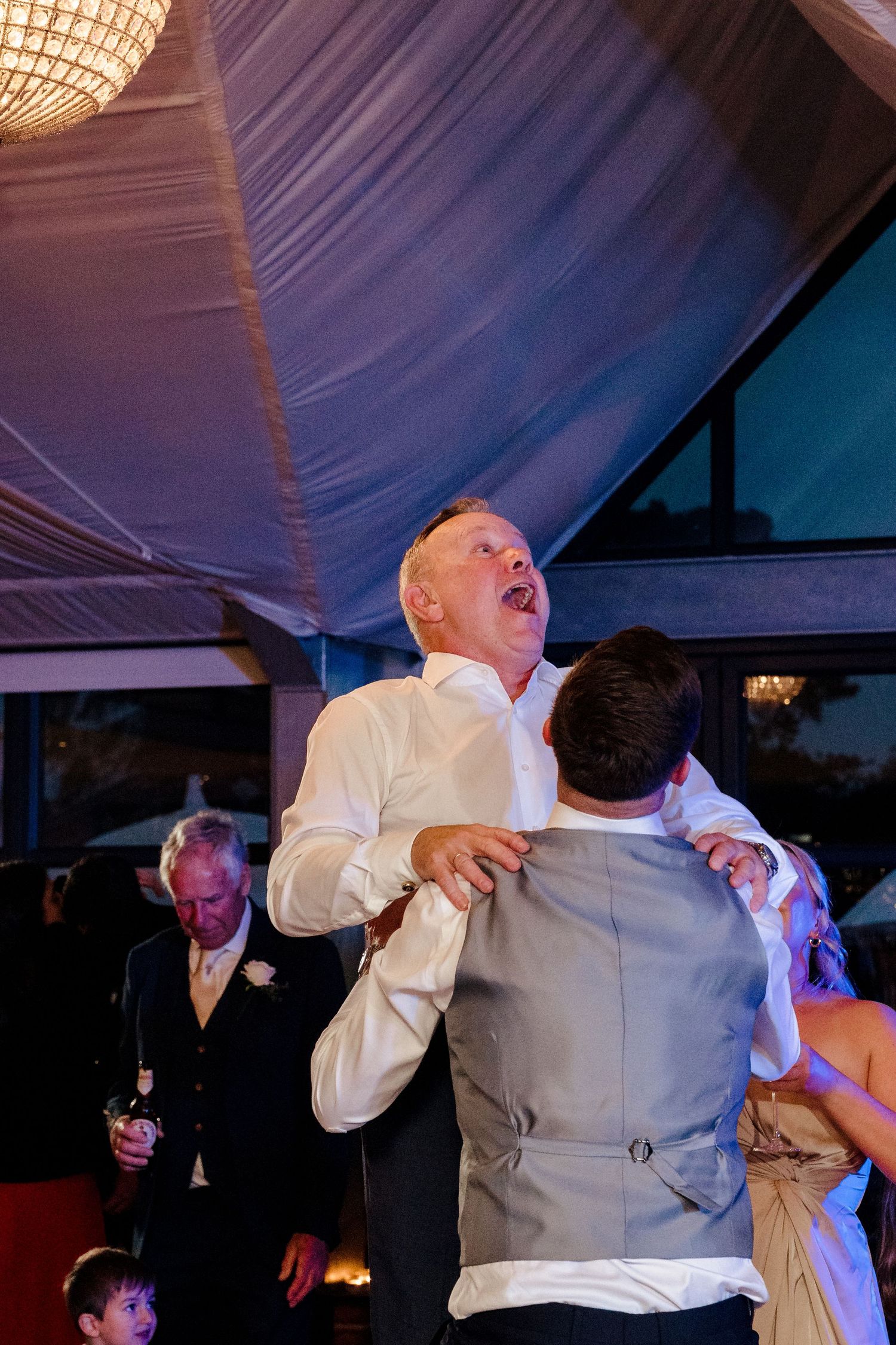 Excited wedding guest being lifted by groom during lively wedding reception dance floor celebration.