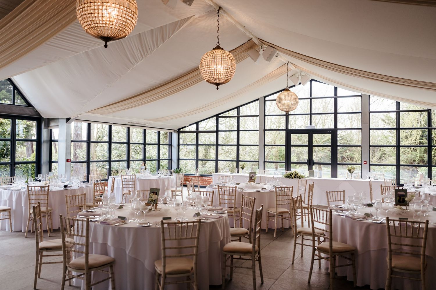 Elegant wedding reception hall with draped ceiling, gold chiavari chairs, round tables and large windows.
