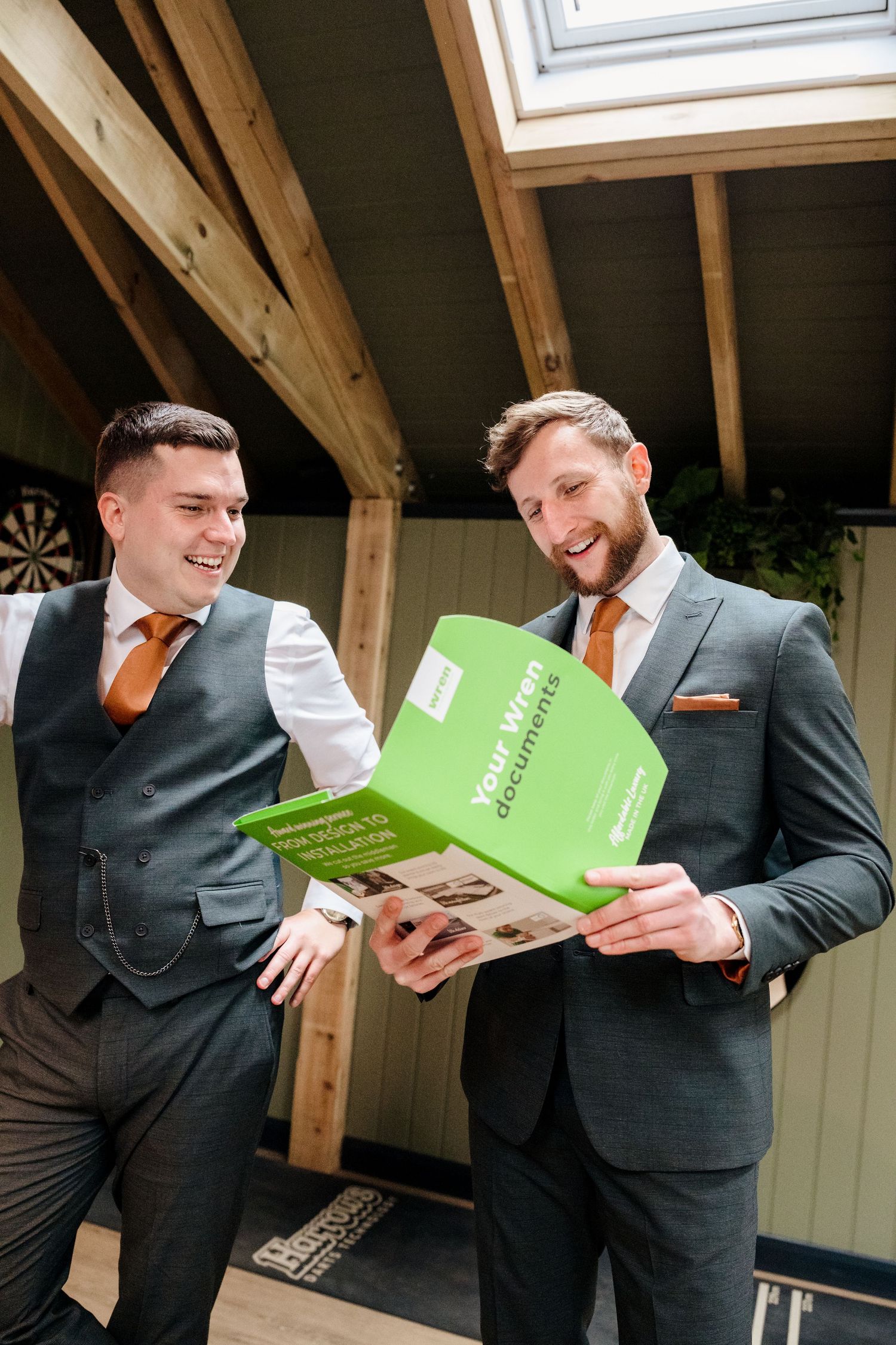 Two grooms in grey suits with orange ties laugh while reviewing wedding documents together.