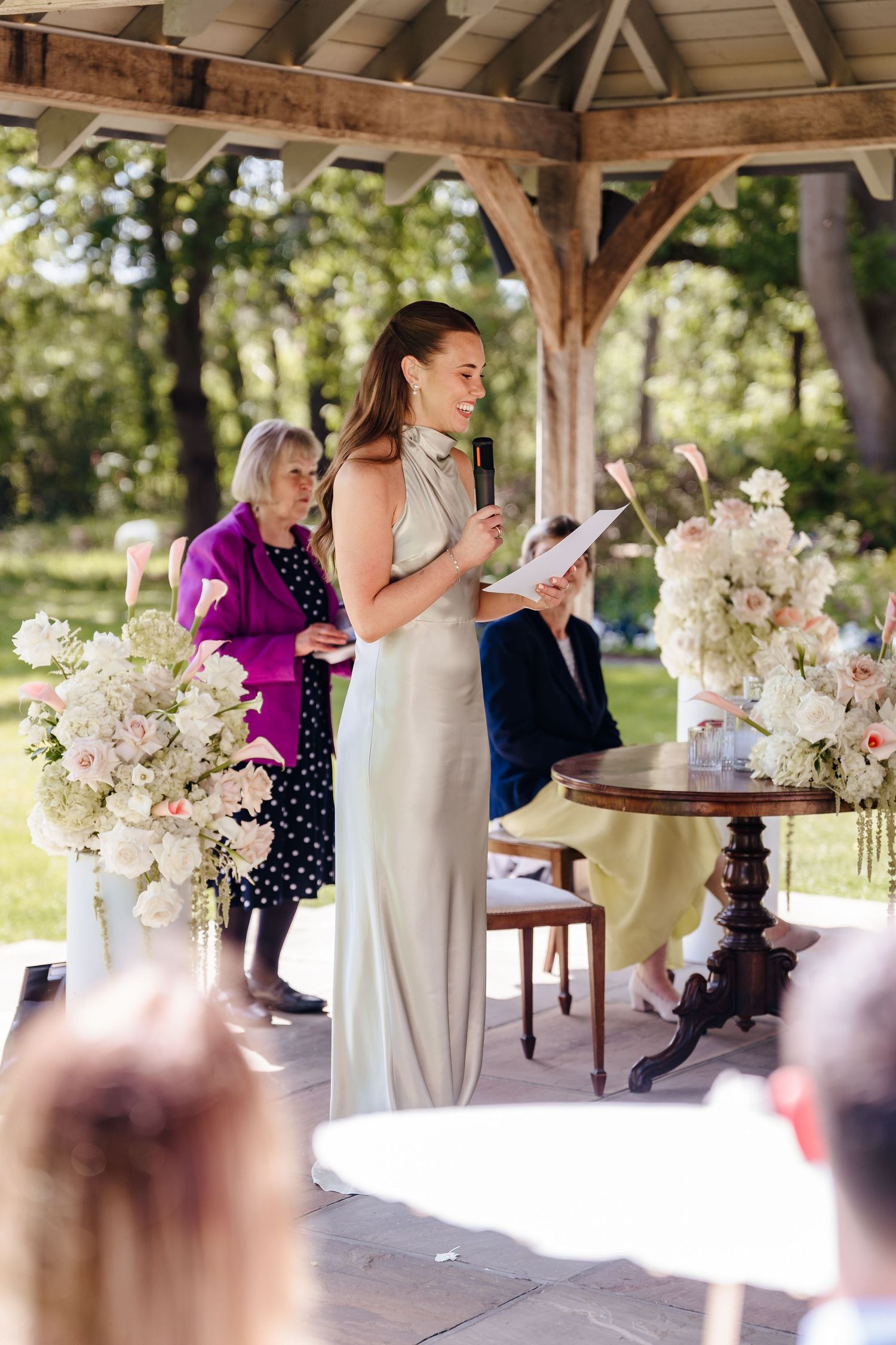 Bridesmaid in a sage green dress reading a speech at an outdoor wedding ceremony.