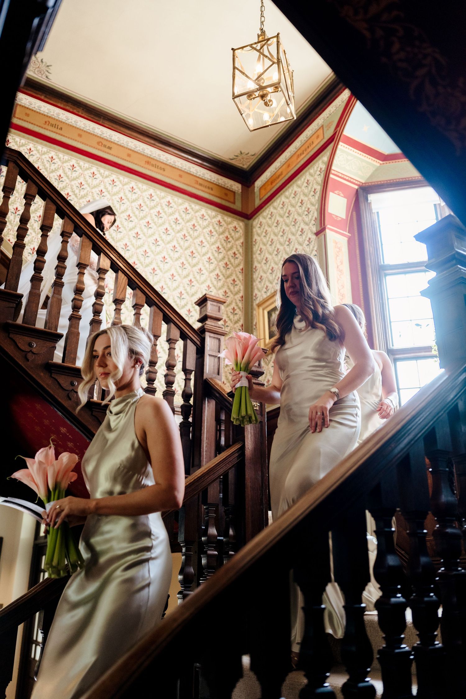 Bride and bridesmaid descending ornate wooden staircase in elegant venue holding pink tulip bouquets.