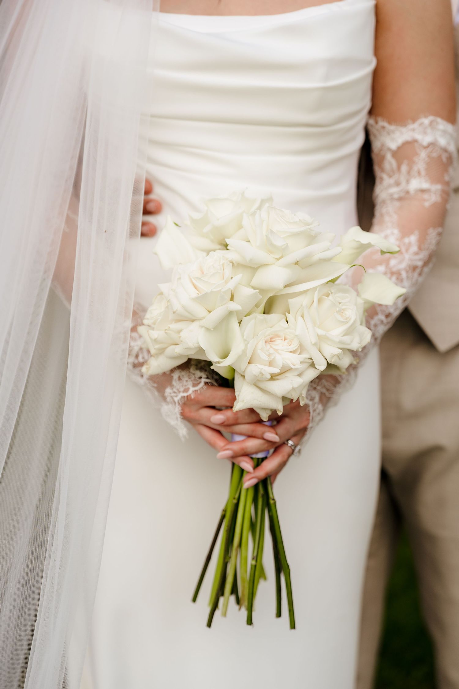 Bride in white gown holds bouquet of white calla lilies and roses with groom beside her.