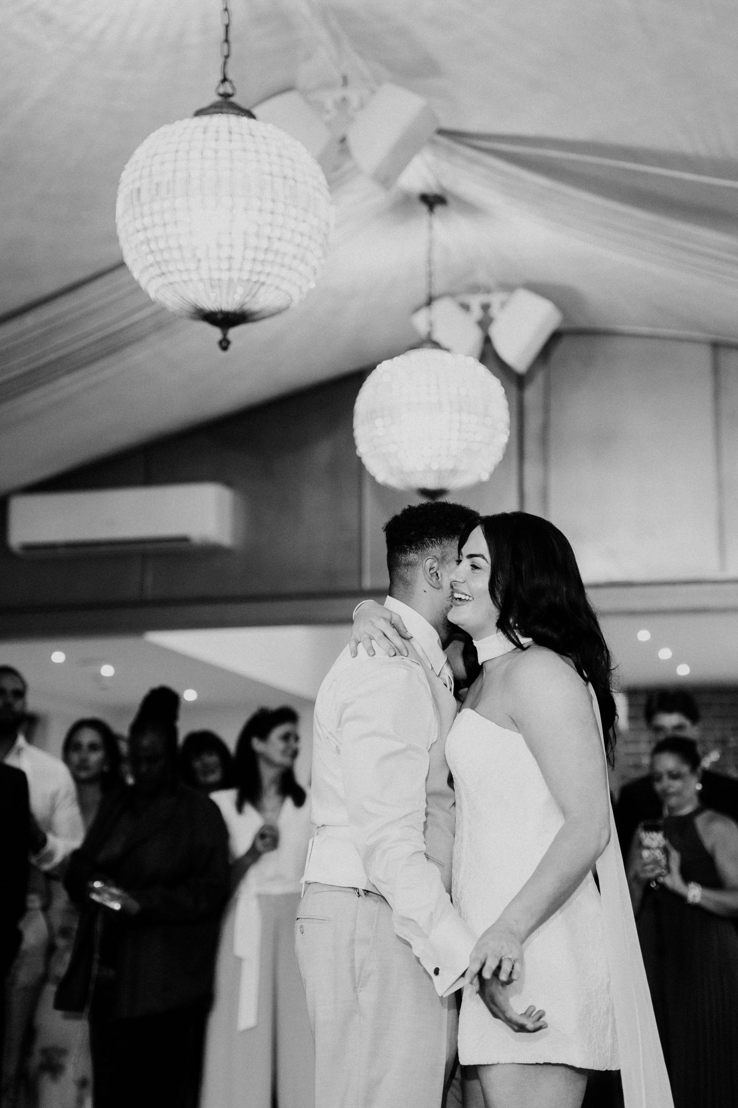 Black and white photo of bride and groom sharing intimate first dance under globe chandeliers.