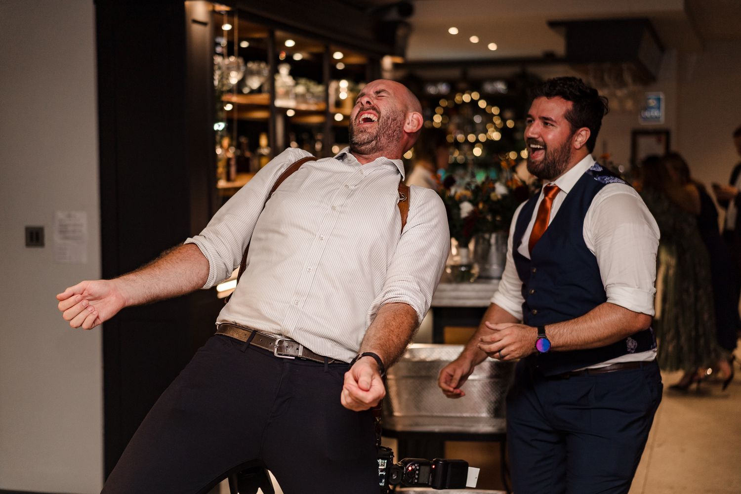 Two men dancing and laughing joyfully at a bar celebration, one in white shirt with suspenders.