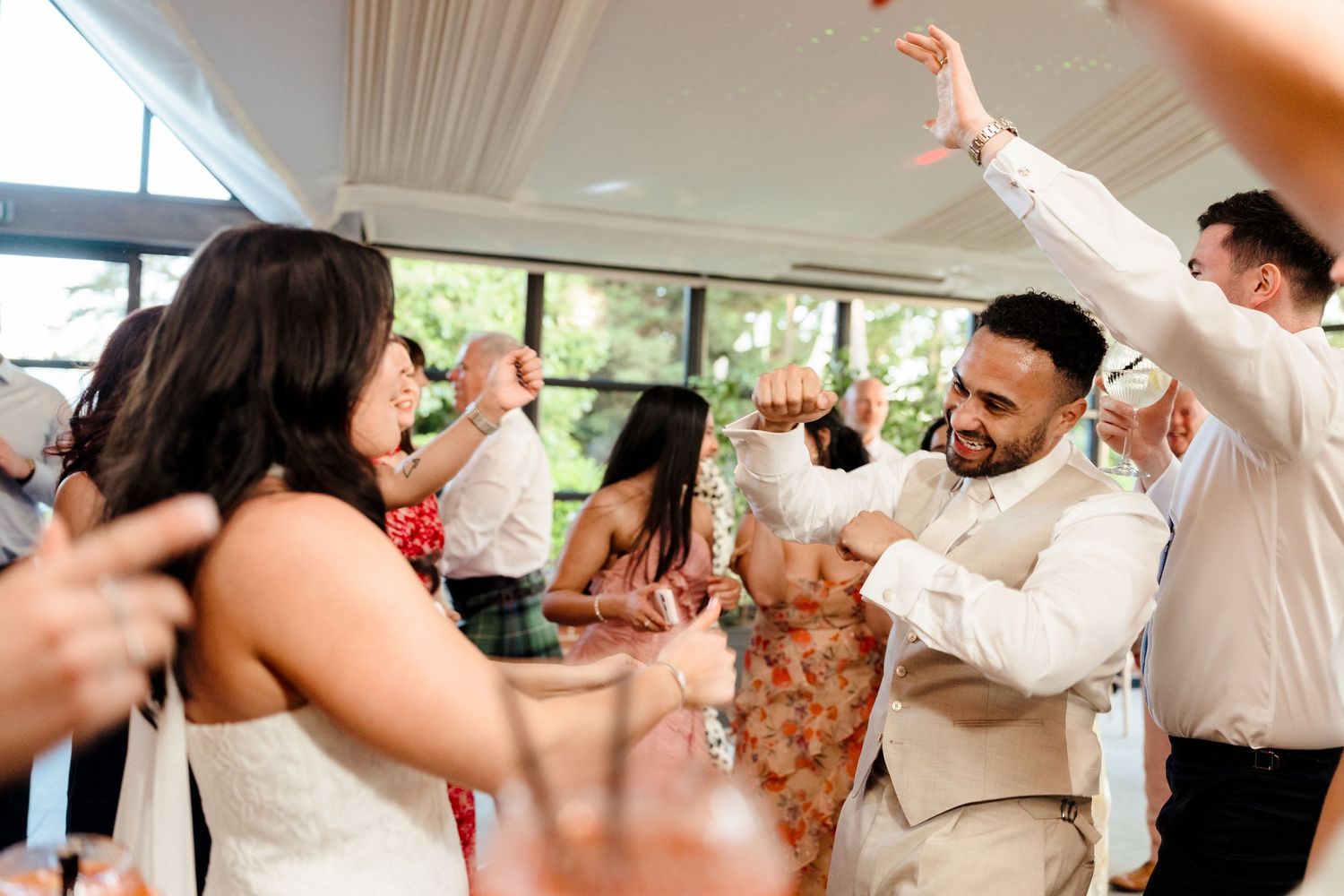 Groom in beige suit dances joyfully with guests at lively wedding reception celebration.