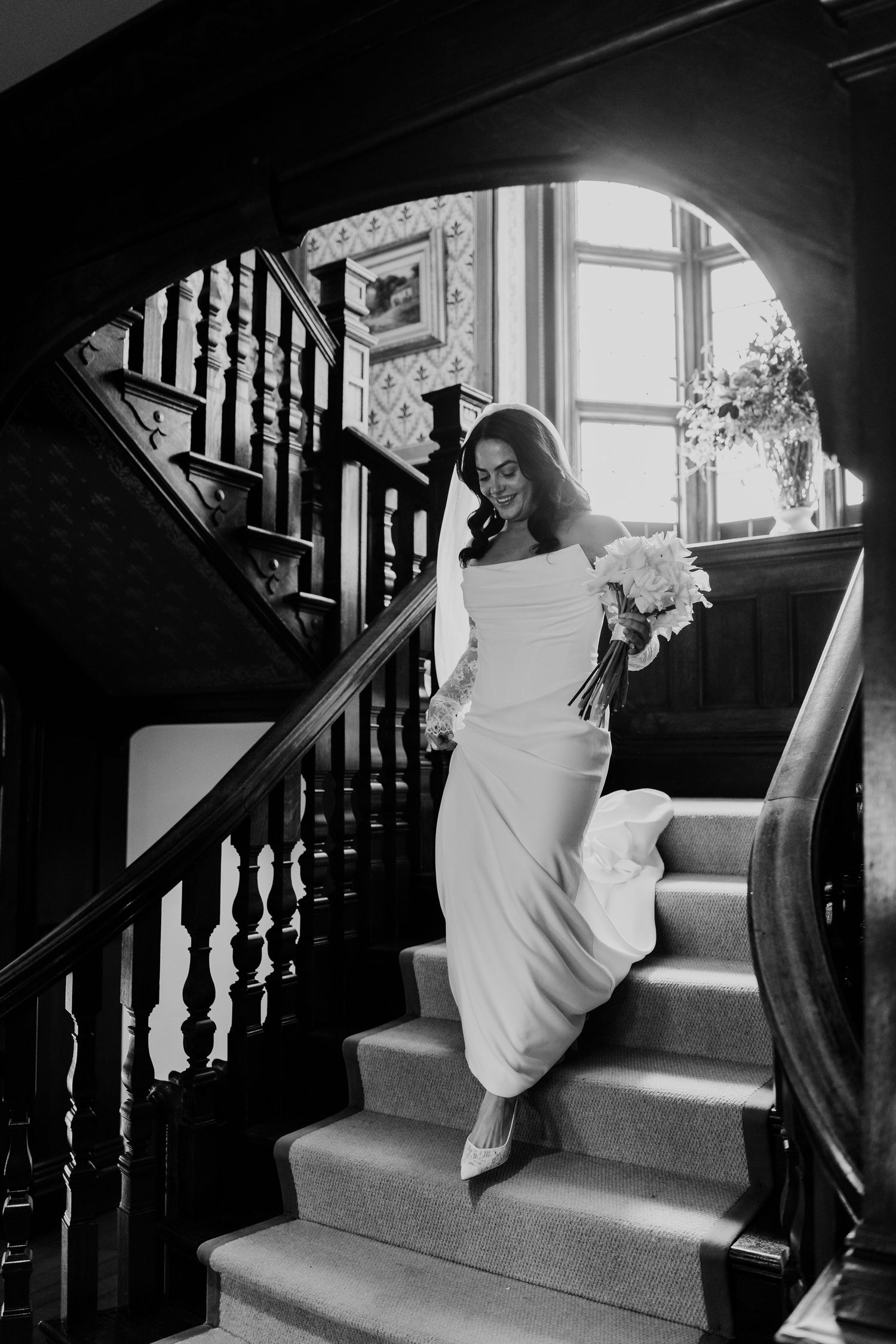 Black and white photo of a bride in a white gown descending a grand wooden staircase.