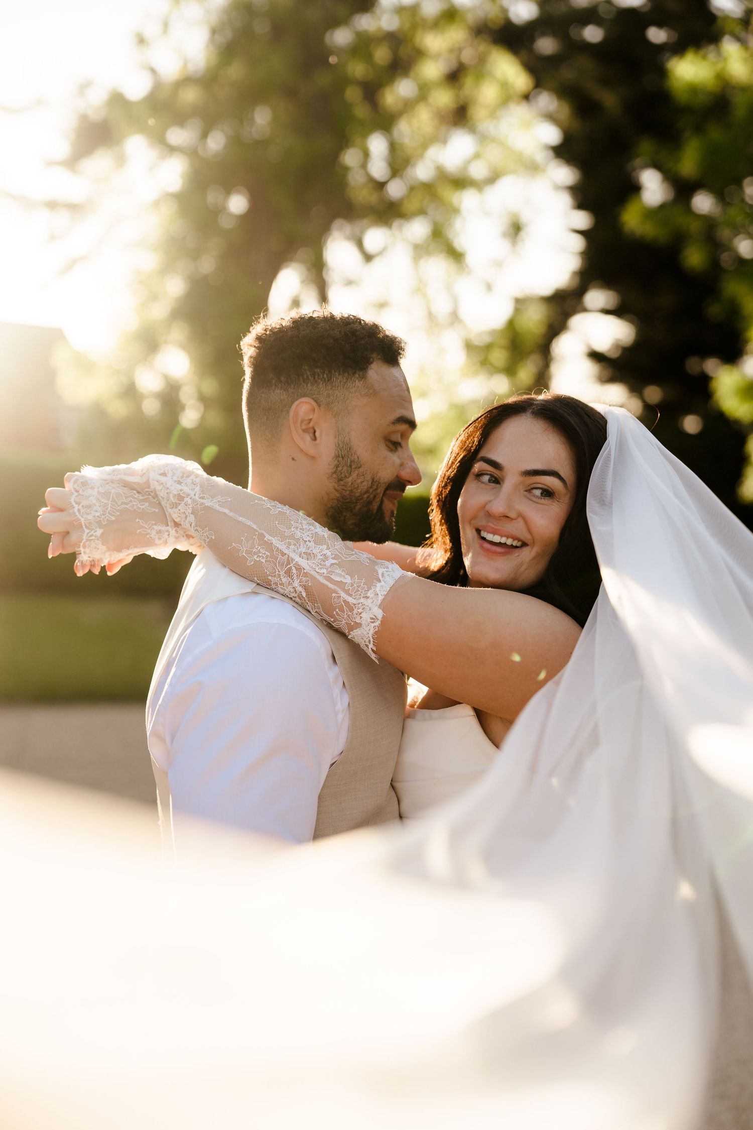 Bride and groom embracing in golden sunlight with veil flowing in breeze.
