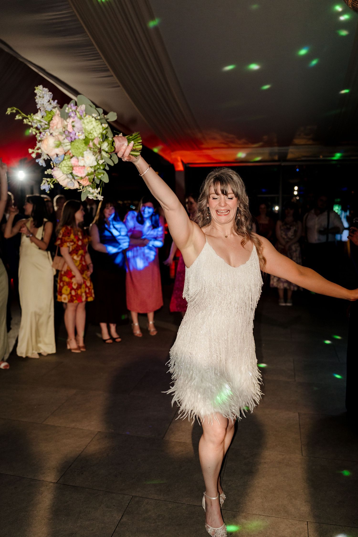 Bride in white feather fringe dress triumphantly holding a pastel floral bouquet on the wedding reception dance floor.
