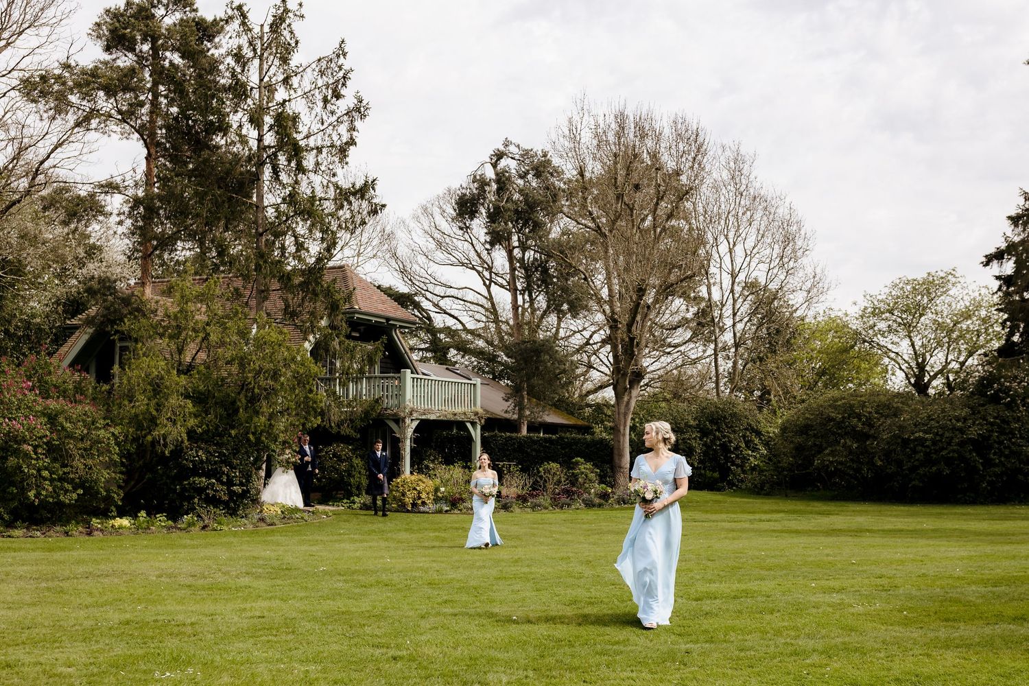 Bridesmaids in light blue dresses walking across a green lawn toward a charming country house venue.