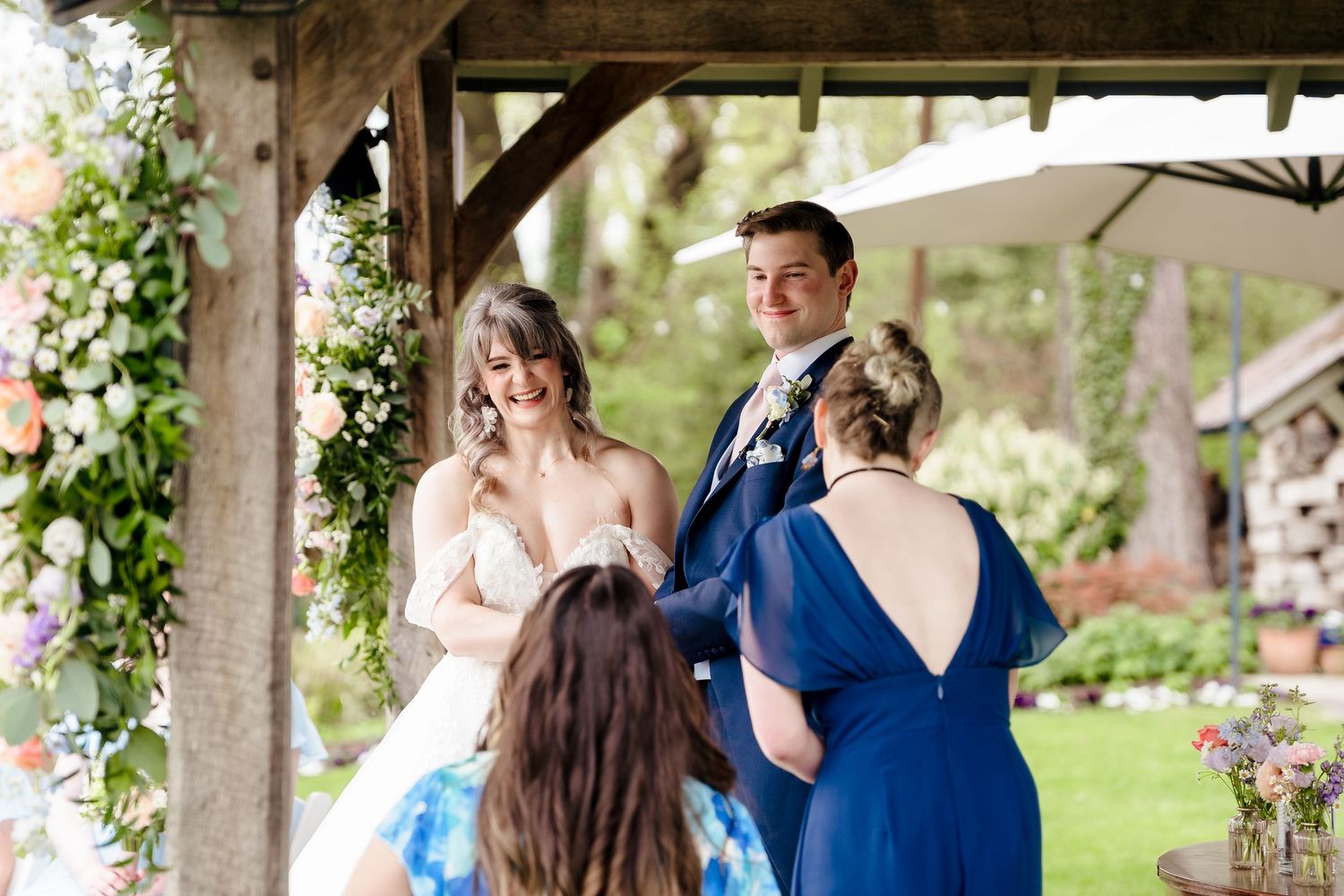 Bride and groom smiling during outdoor wedding ceremony under floral arch, officiant in blue dress.