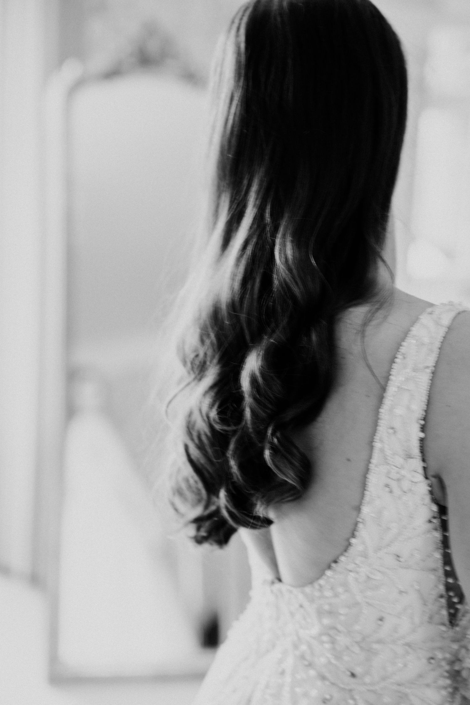 Black and white photo of bride with long curly hair in a backless beaded wedding gown facing a mirror.