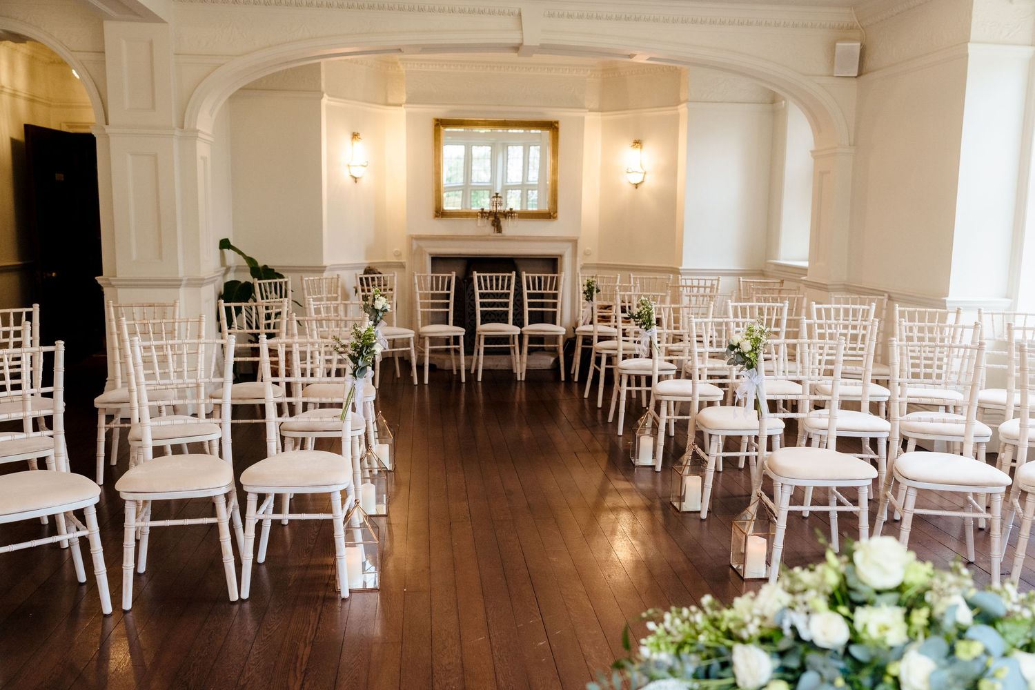 Elegant wedding ceremony room with white Chiavari chairs arranged in rows on hardwood floors, adorned with floral decorations.