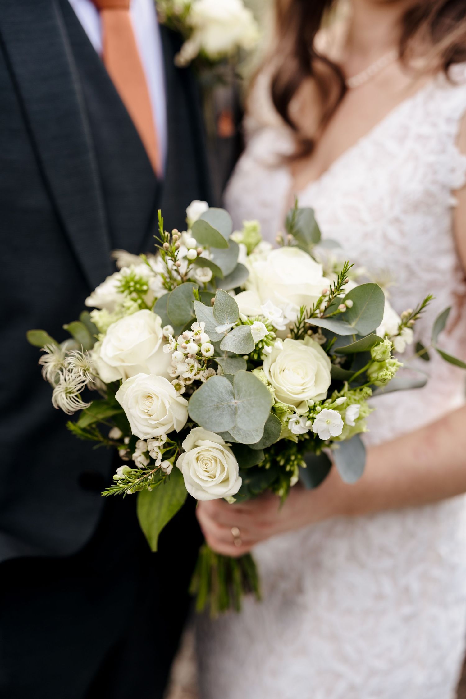 Bride holding elegant white rose and eucalyptus wedding bouquet beside groom in dark suit.