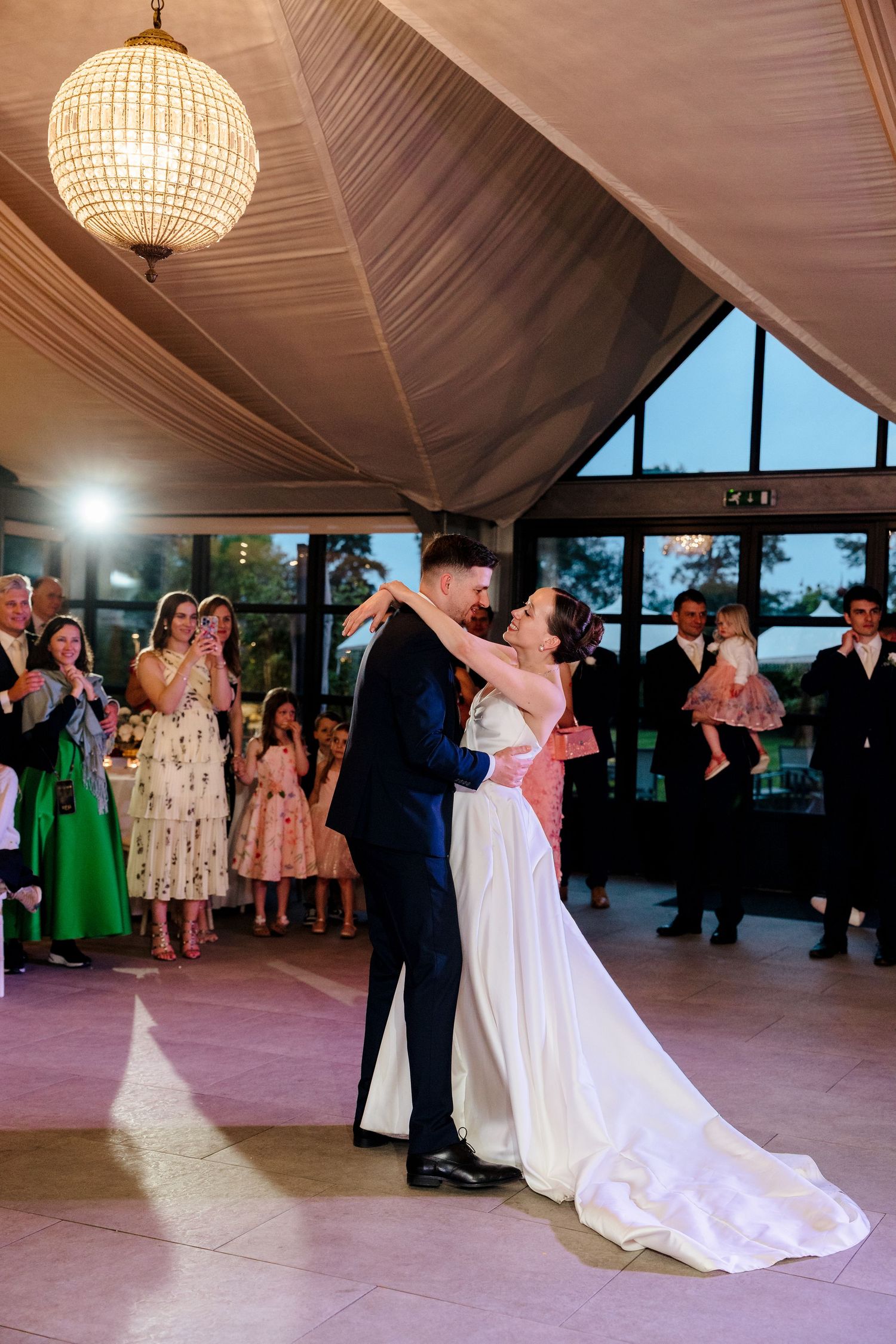 Bride and groom share first dance under chandelier while wedding guests watch in elegant reception venue.