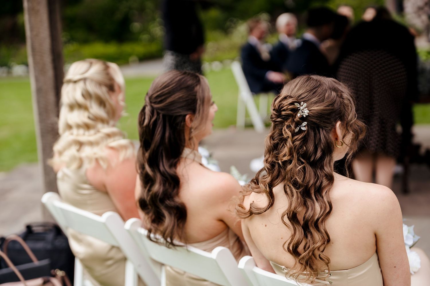 Back view of bridesmaids with elegant curled hairstyles seated at outdoor wedding ceremony.