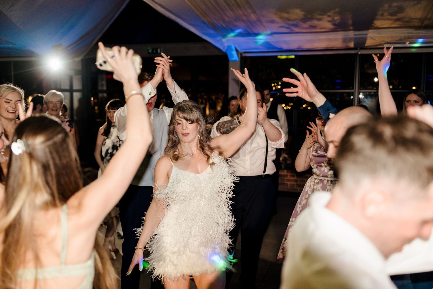 Bride in white feather fringe dress dancing with raised hands among wedding guests on a lit reception dance floor.