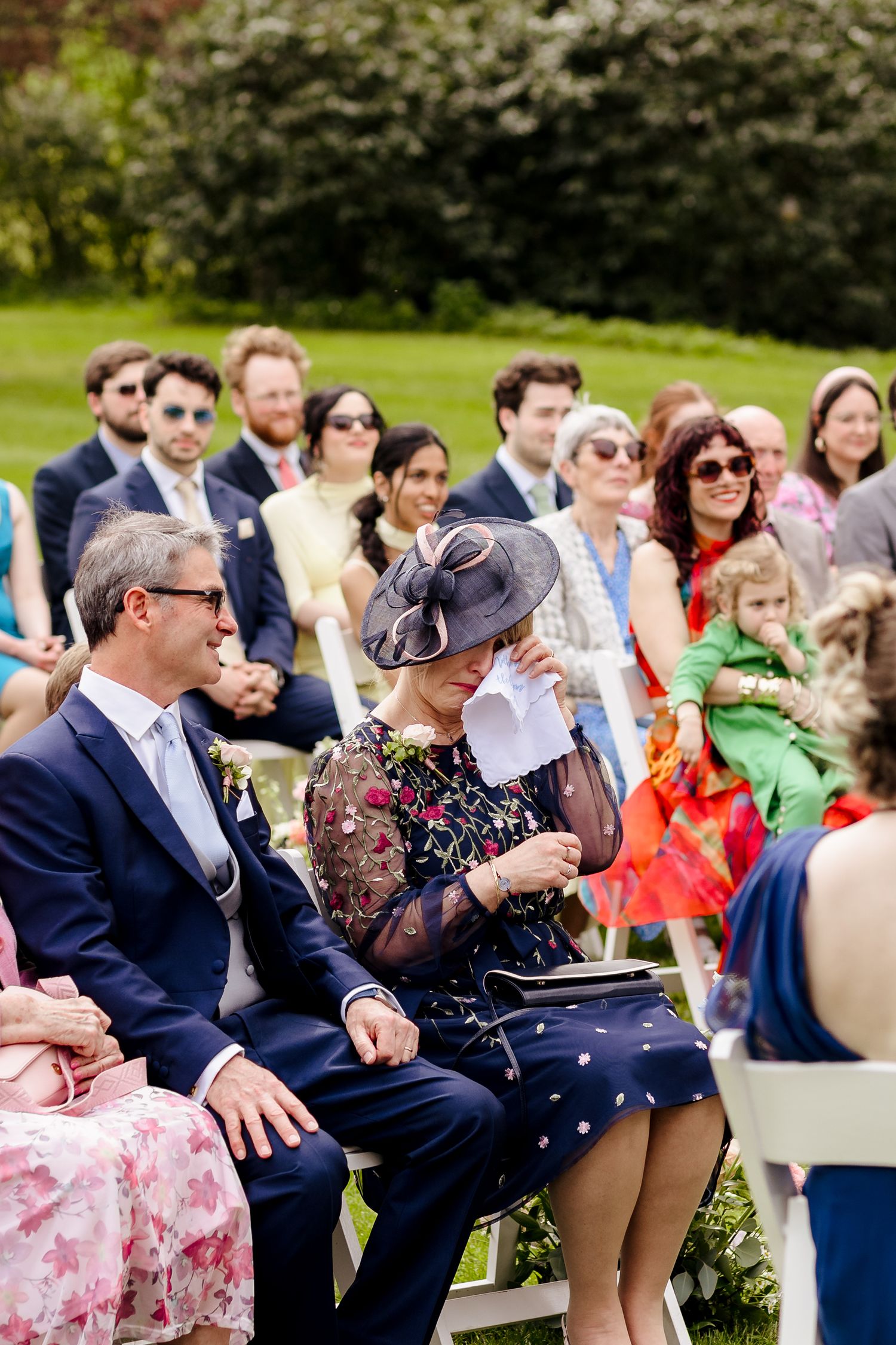 Emotional wedding guest in floral dress dabs eyes with tissue while seated at outdoor ceremony.