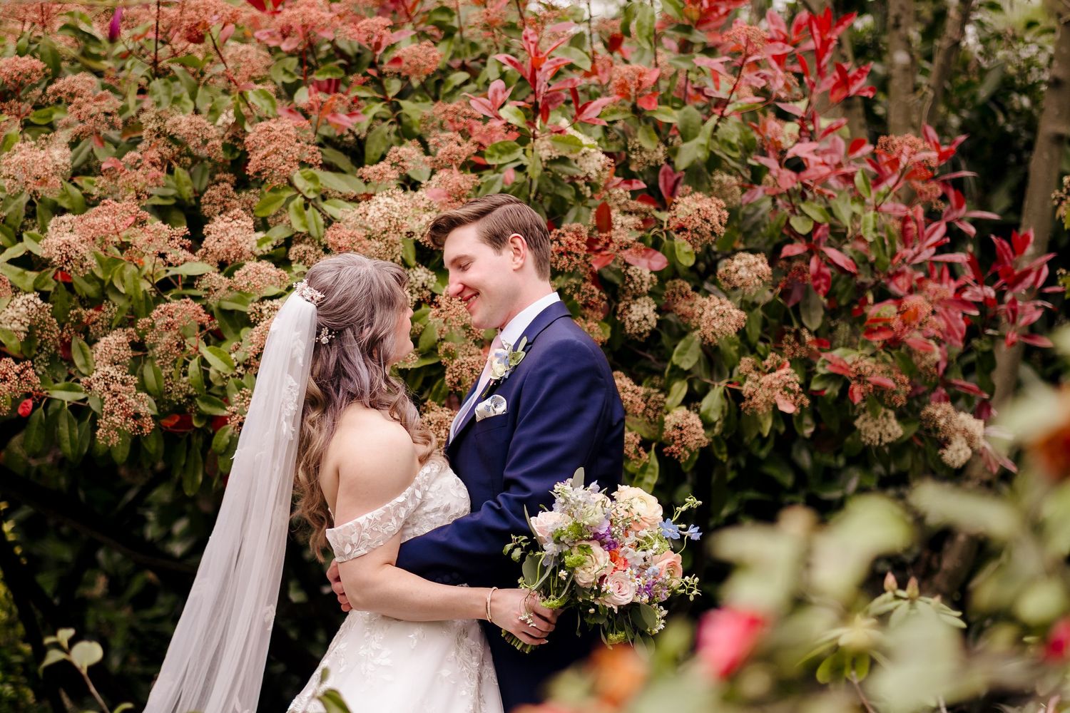 Bride and groom smiling at each other in front of vibrant red and orange floral bushes outdoors.