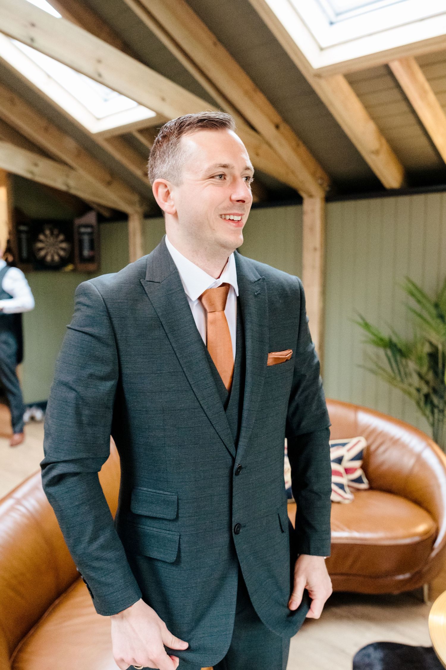 Smiling groom in dark green three-piece suit with burnt orange tie and pocket square at wedding venue.