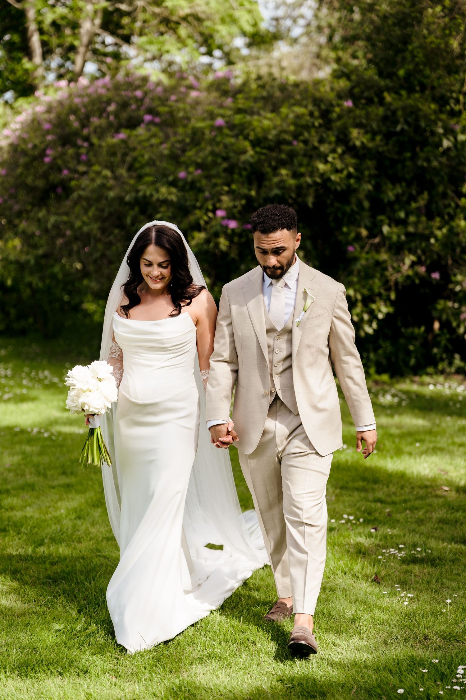 Bride and groom in beige suit walk hand in hand across green lawn surrounded by blooming garden.