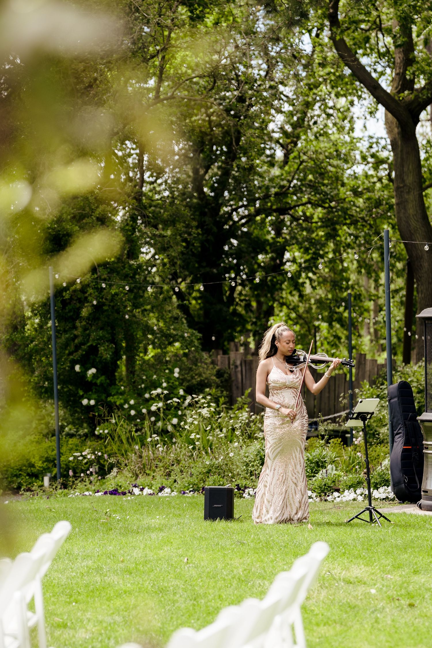A violinist in a gold gown performs outdoors at a garden wedding ceremony surrounded by lush green trees.